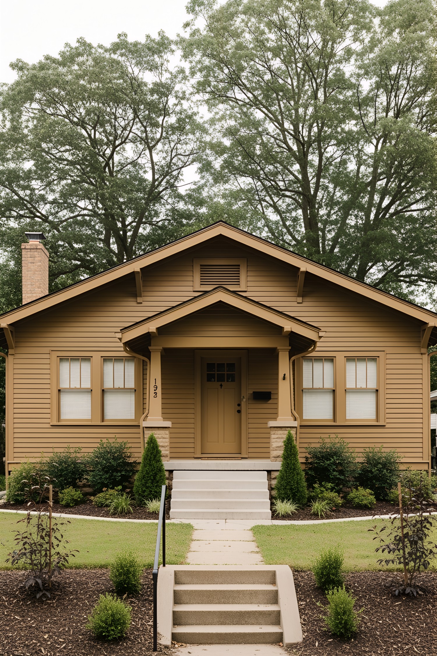 1930s bungalow, symmetrical single-story facade, horizontal wood siding, rectangular footprint with a centered front porch projection, gabled roof with moderate overhang and exposed rafter tails, paired double-hung windows with simple trim, solid single front door with a small covered stoop supported by wood columns, narrow concrete steps leading to porch, modest lawn bordered by mature shrubs and small trees, informal landscaping, residential neighborhood setting with large deciduous trees in the background.