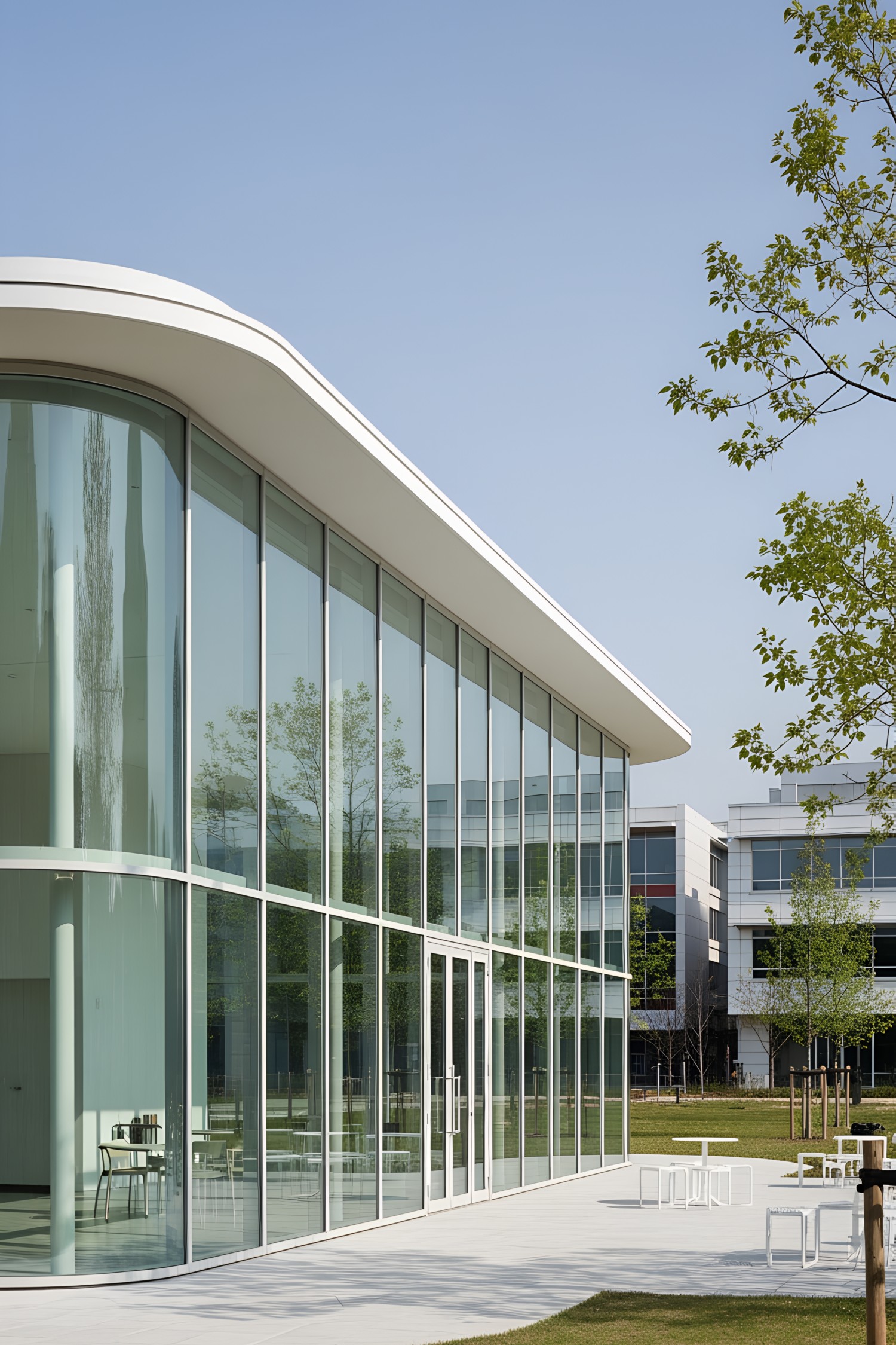 Modern glass pavilion with transparent walls and a curved roof, surrounded by greenery
