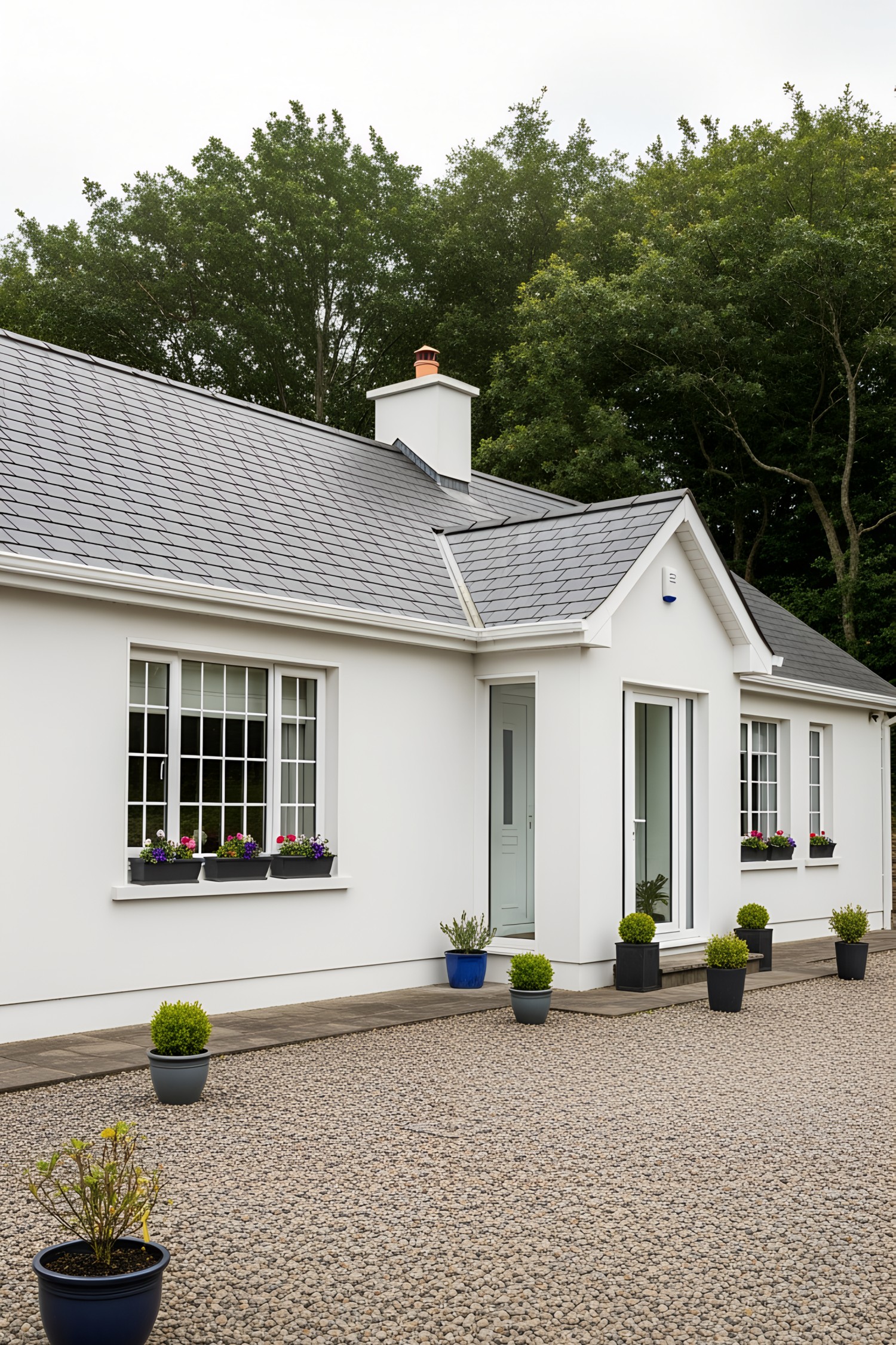 White bungalow with gray roof and potted plants, surrounded by trees