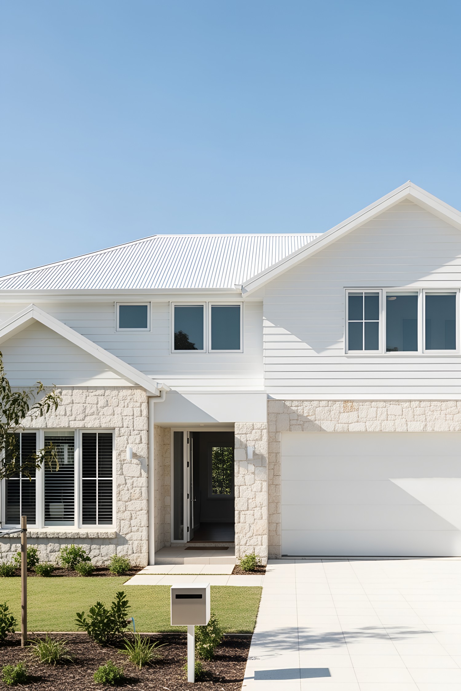 Modern coastal facade, single-story structure with clean lines, white horizontal siding, light-colored natural stone feature elements, wide gabled roof with white corrugated panels, large rectangular windows with white framing, prominent white garage door, integrated entrance with minimal trim, smooth white paved driveway, small manicured lawn with sparse planting, bright sunlit setting, clear sky environment.