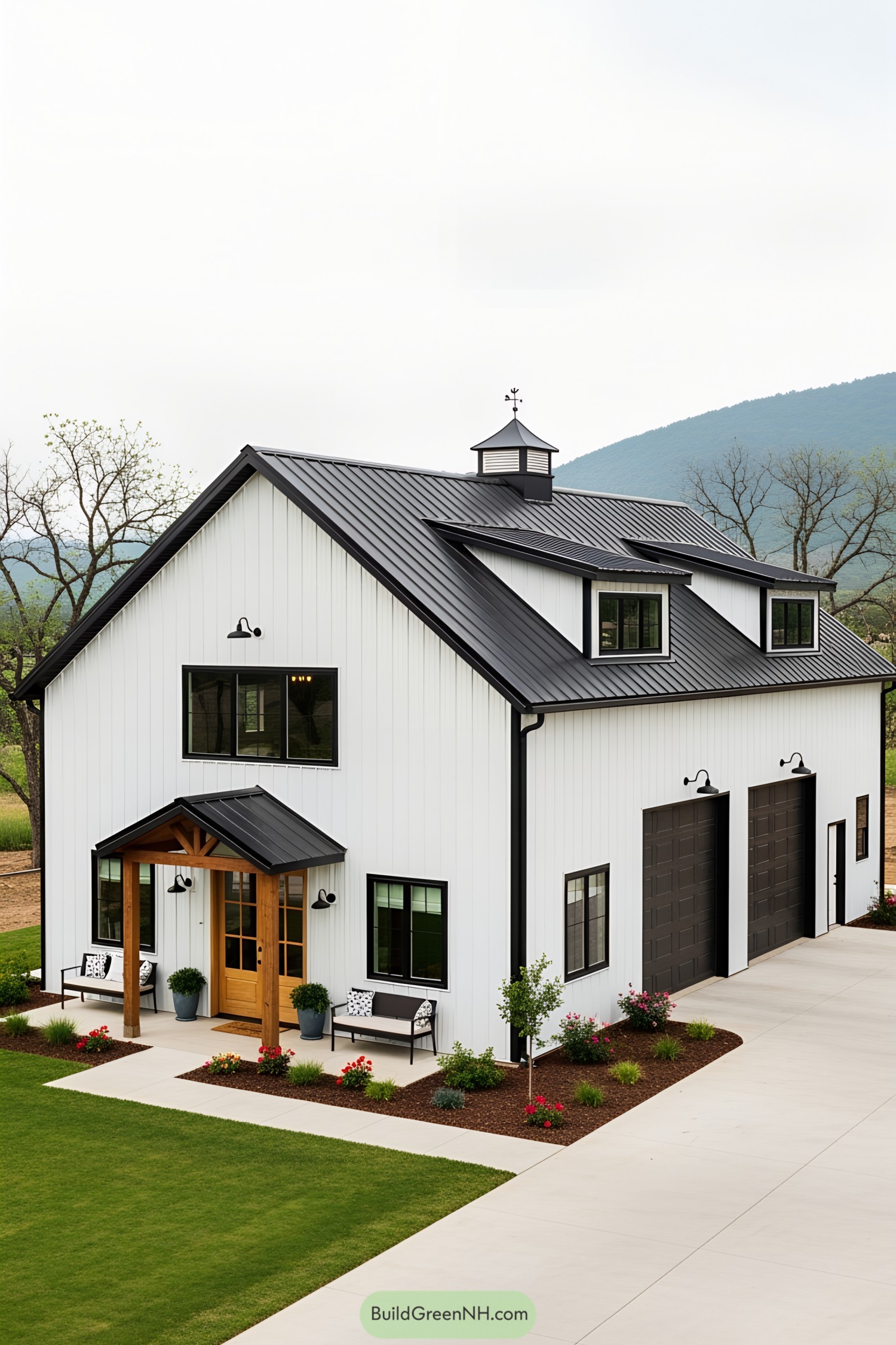 White barndominium with black roof and garage doors, surrounded by greenery