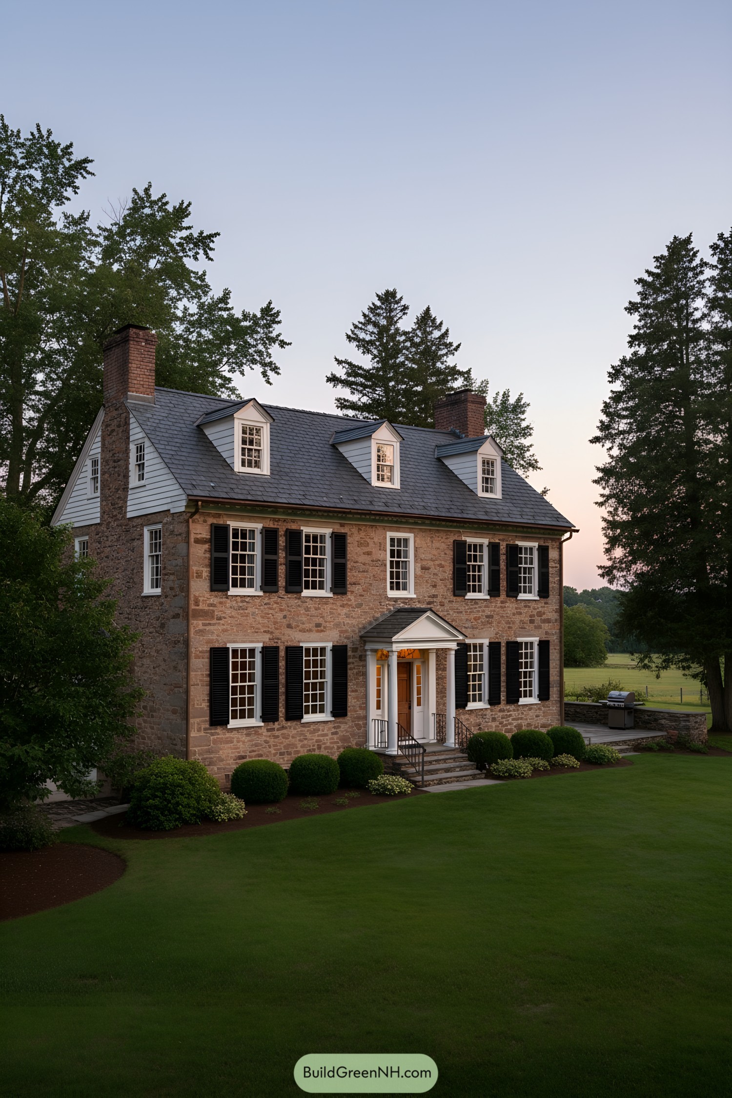 Three-story colonial farmhouse with fieldstone walls, black shutters, and a slate roof punctuated by dormers and twin end chimneys