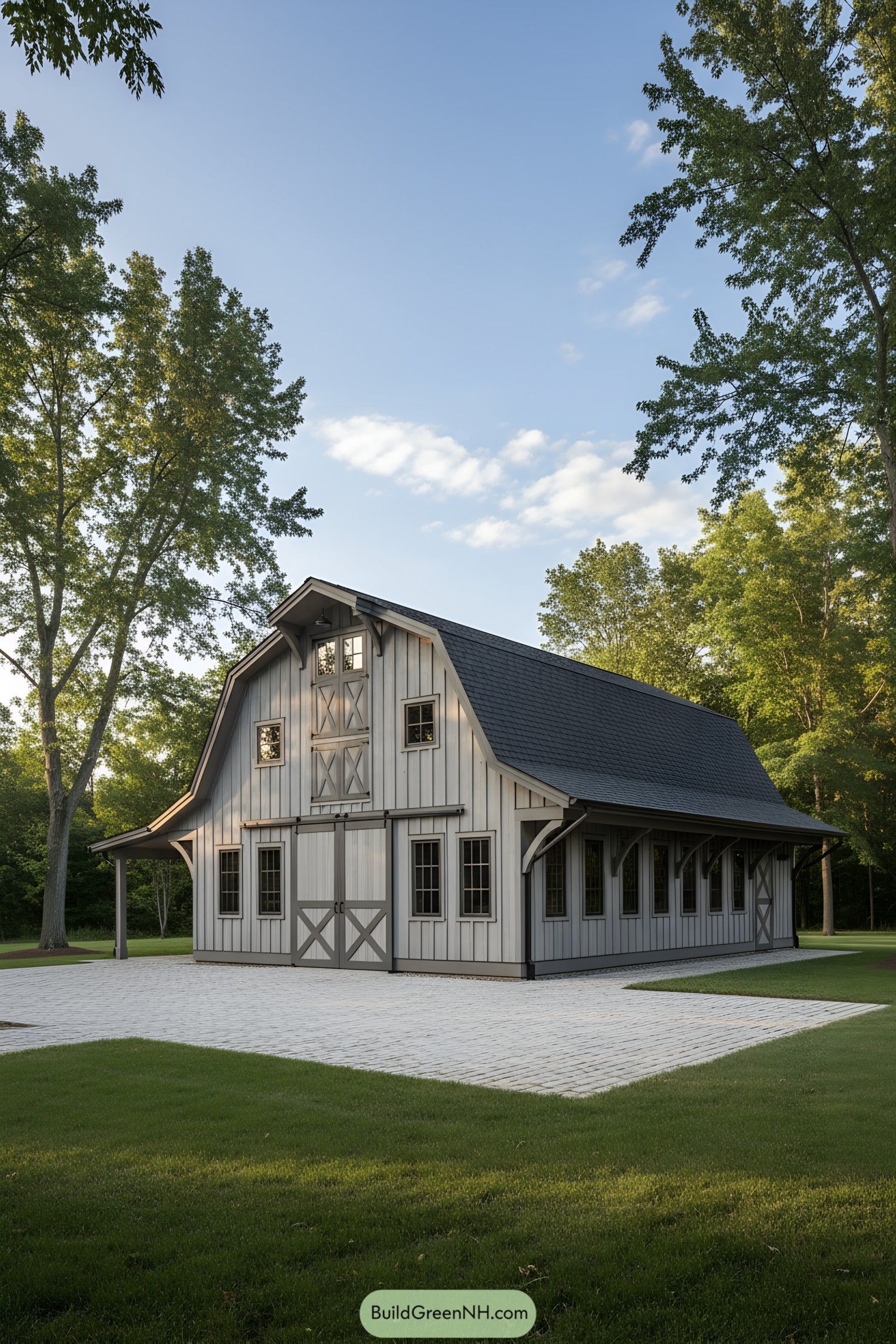 Gray board-and-batten gambrel barn with dormer and crossbuck doors on a paved court, surrounded by tall trees