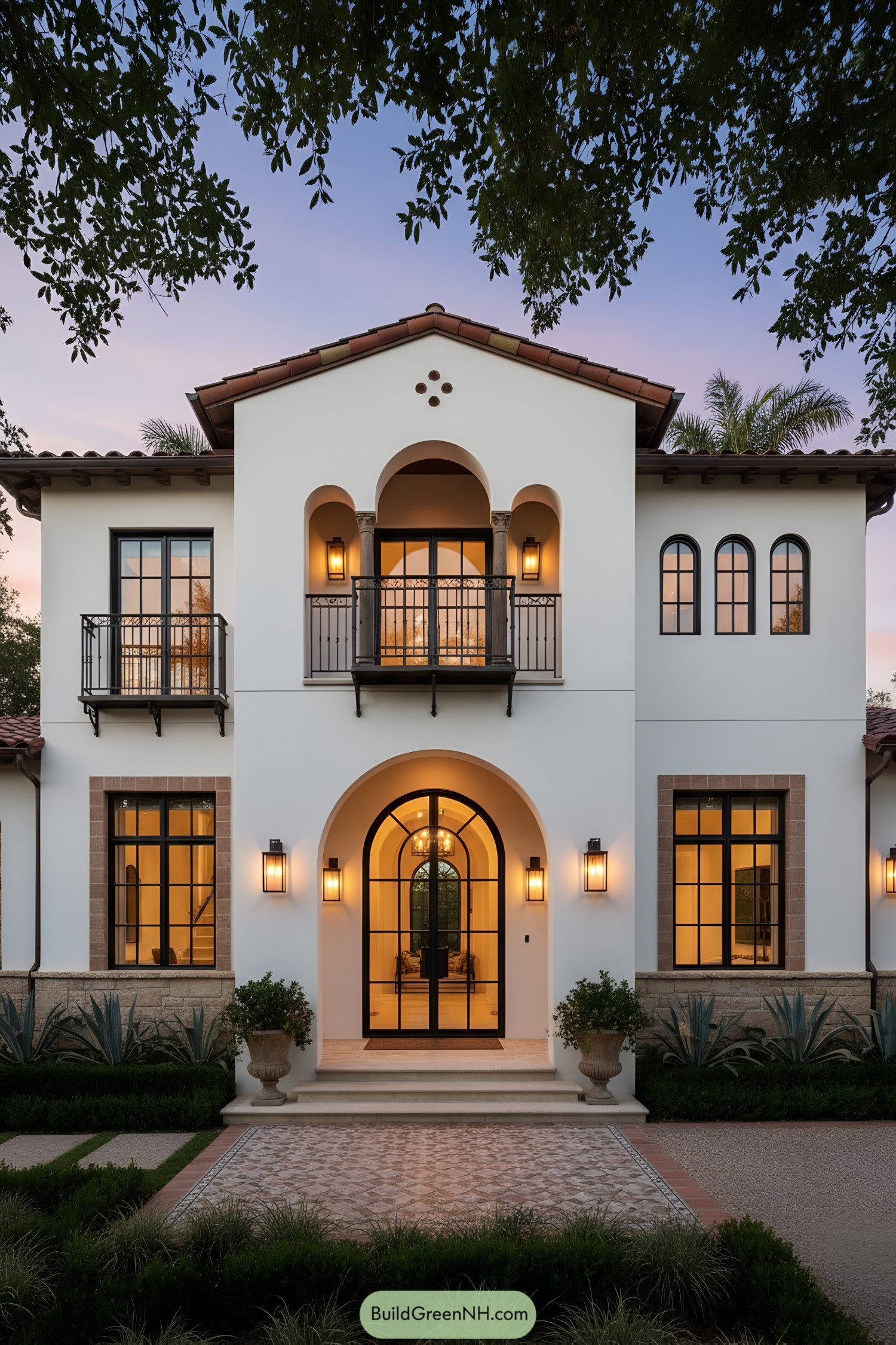 White stucco Mediterranean farmhouse with arched entry, wrought-iron balconies, and warm lantern lighting at dusk