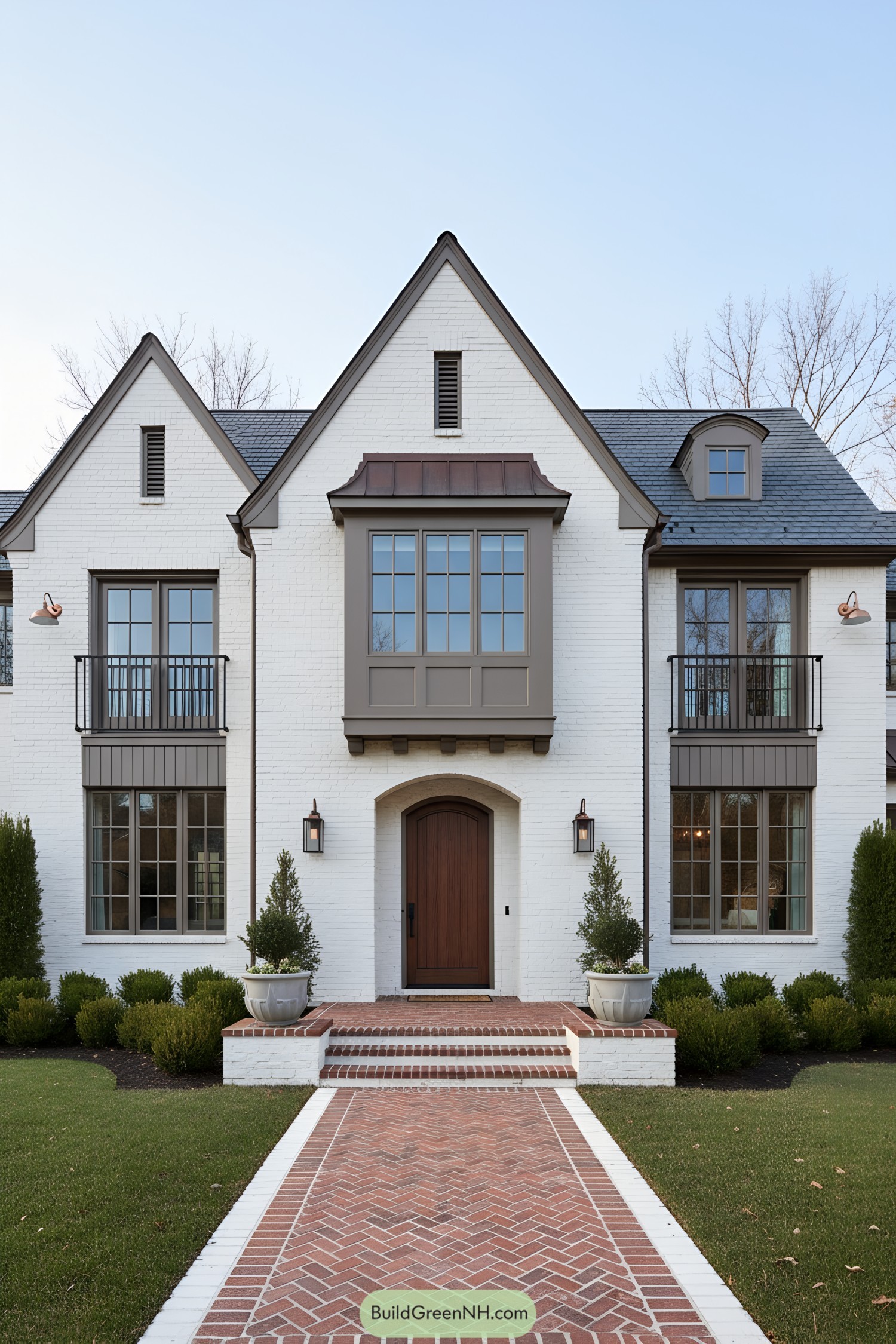 White brick Tudor with copper-capped oriel, arched wood door, slate roof, and twin Juliet balconies