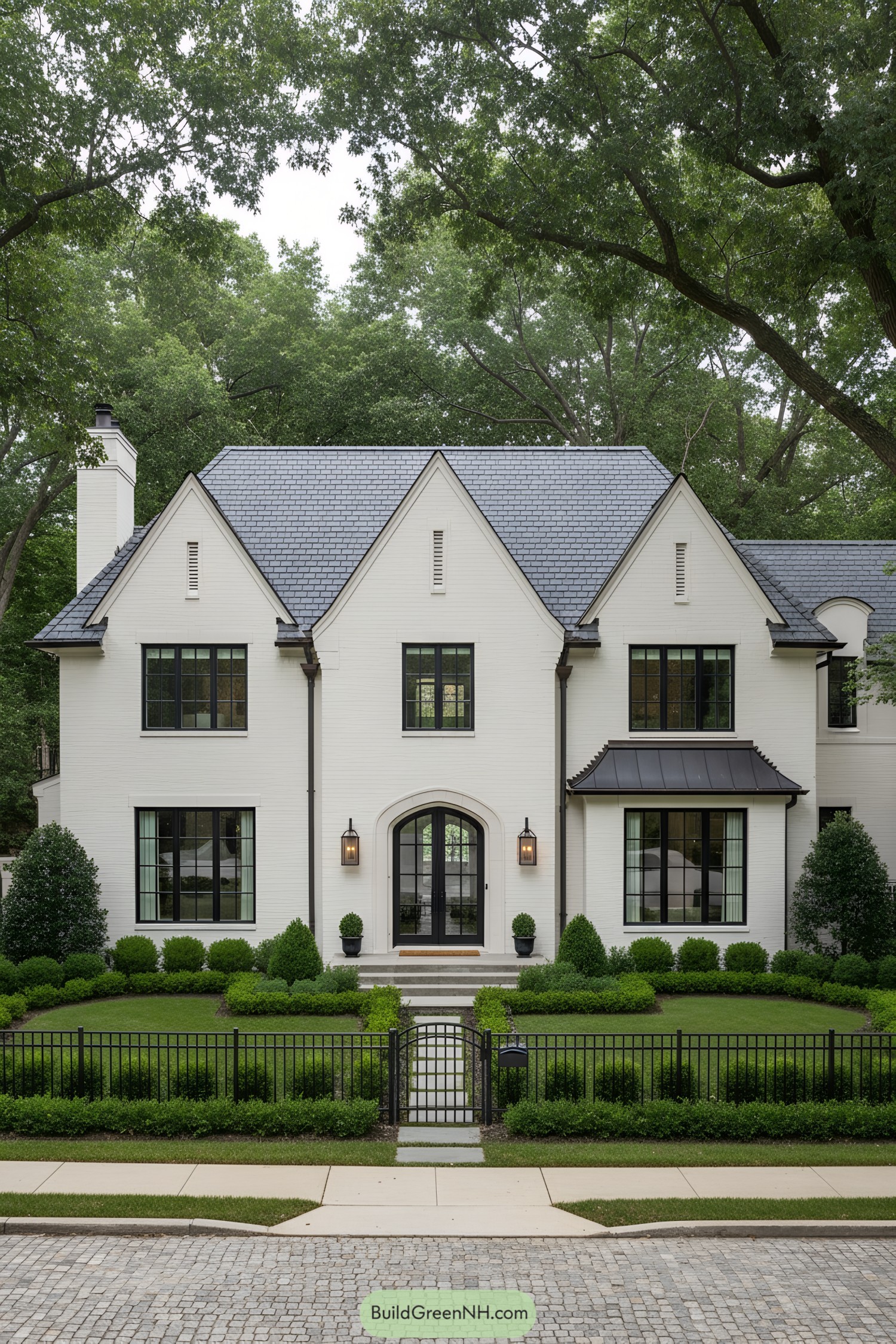 Modern Tudor manor with slate roof, white brick, and black steel windows behind a tidy iron fence and clipped hedges