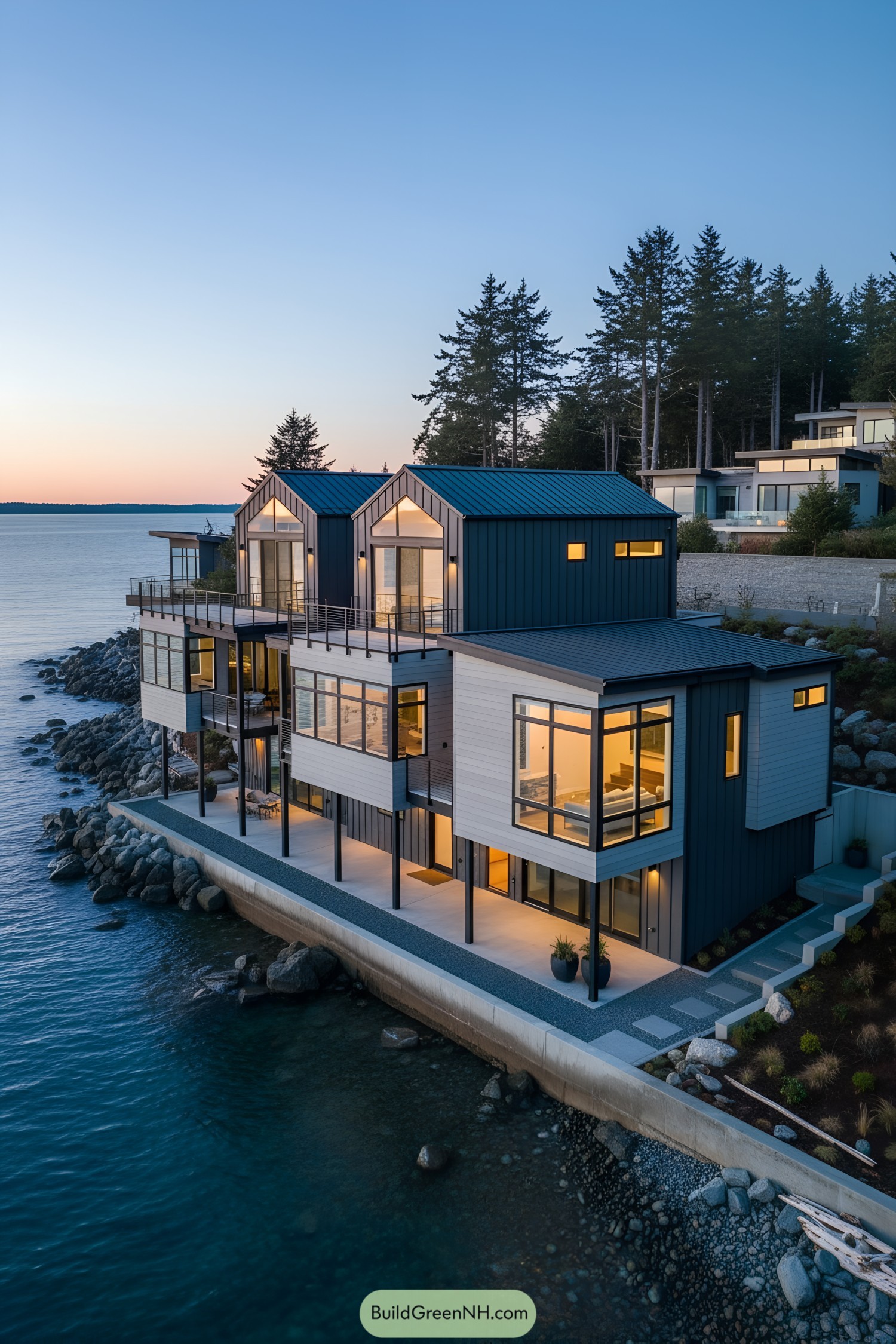 Modern waterfront house with stacked gables and expansive glass perched on a rocky shoreline at dusk