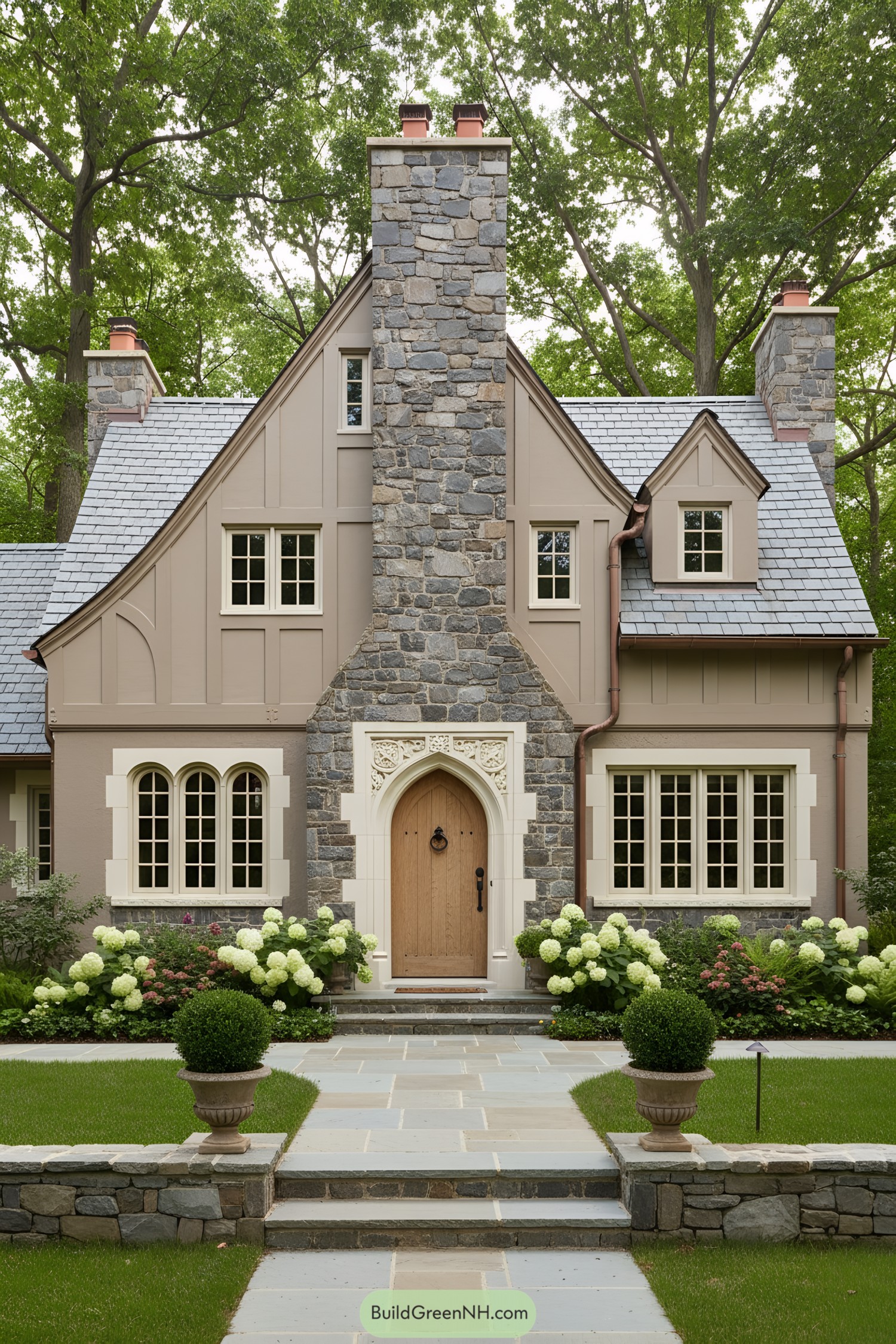 Small Tudor cottage with central stone chimney, arched oak door, slate roof, and trimmed gardens