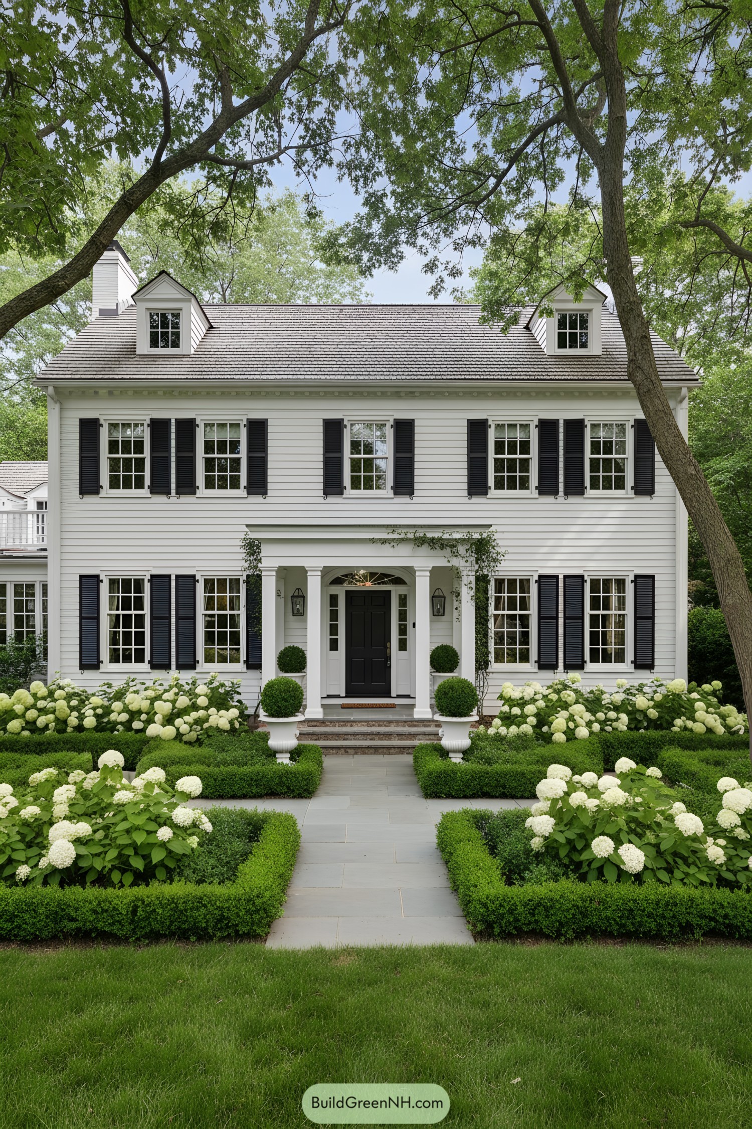White clapboard colonial with black shutters, centered portico, and manicured boxwood gardens