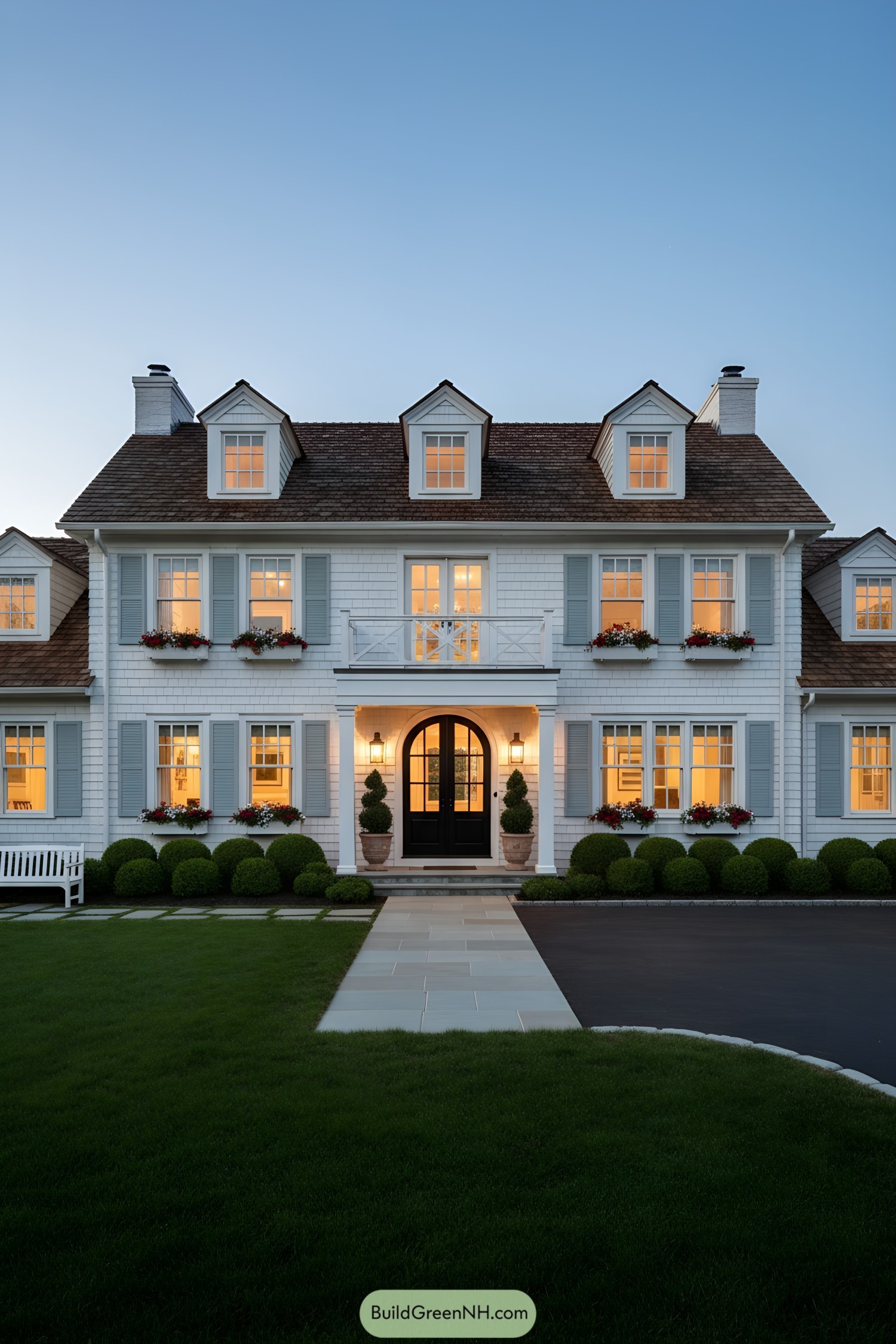 White shingle colonial with blue shutters, three dormers, arched black door, and a small balcony at dusk