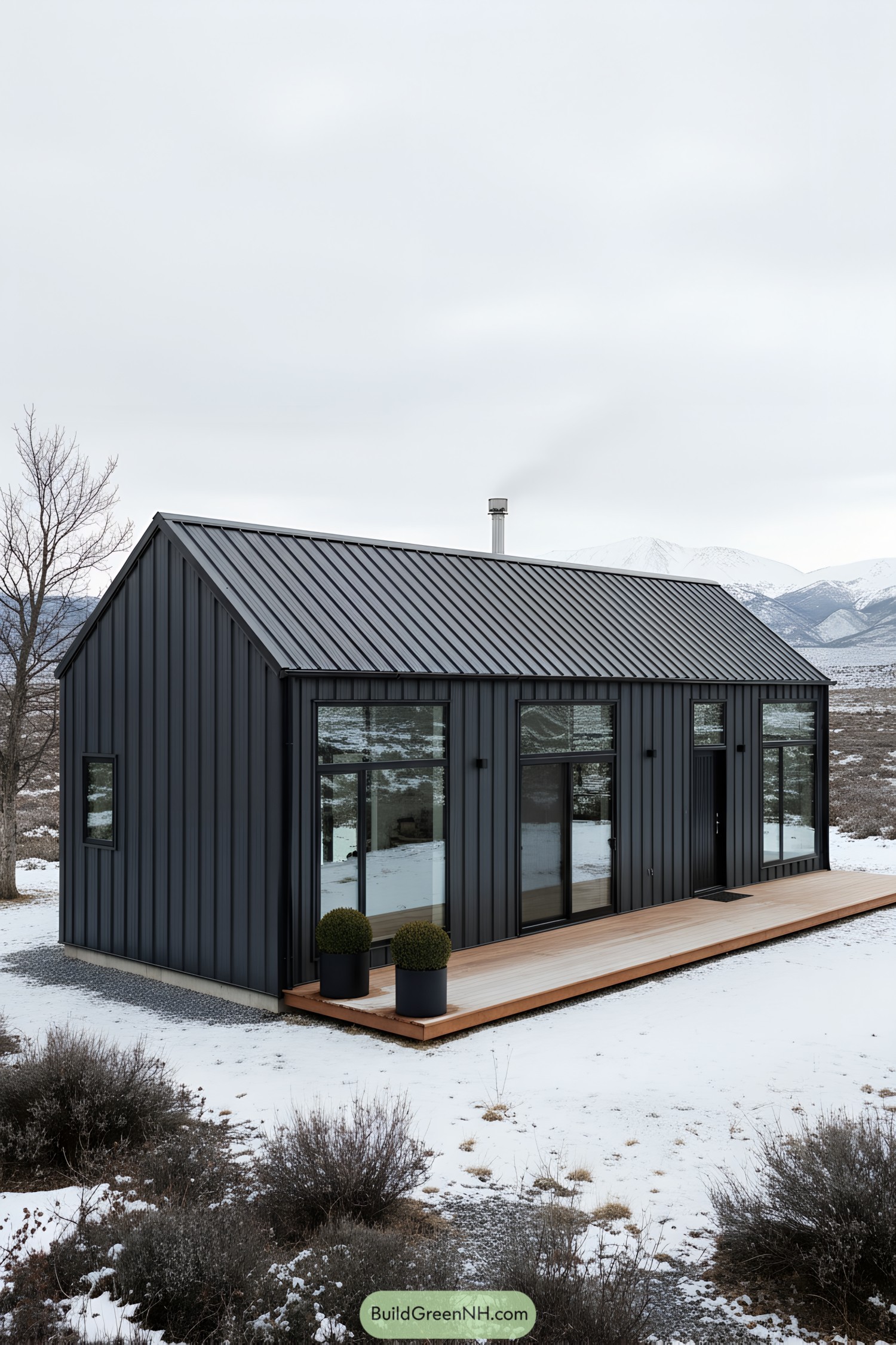 Dark metal gable shed house with tall windows and a slim wood deck in a snowy landscape