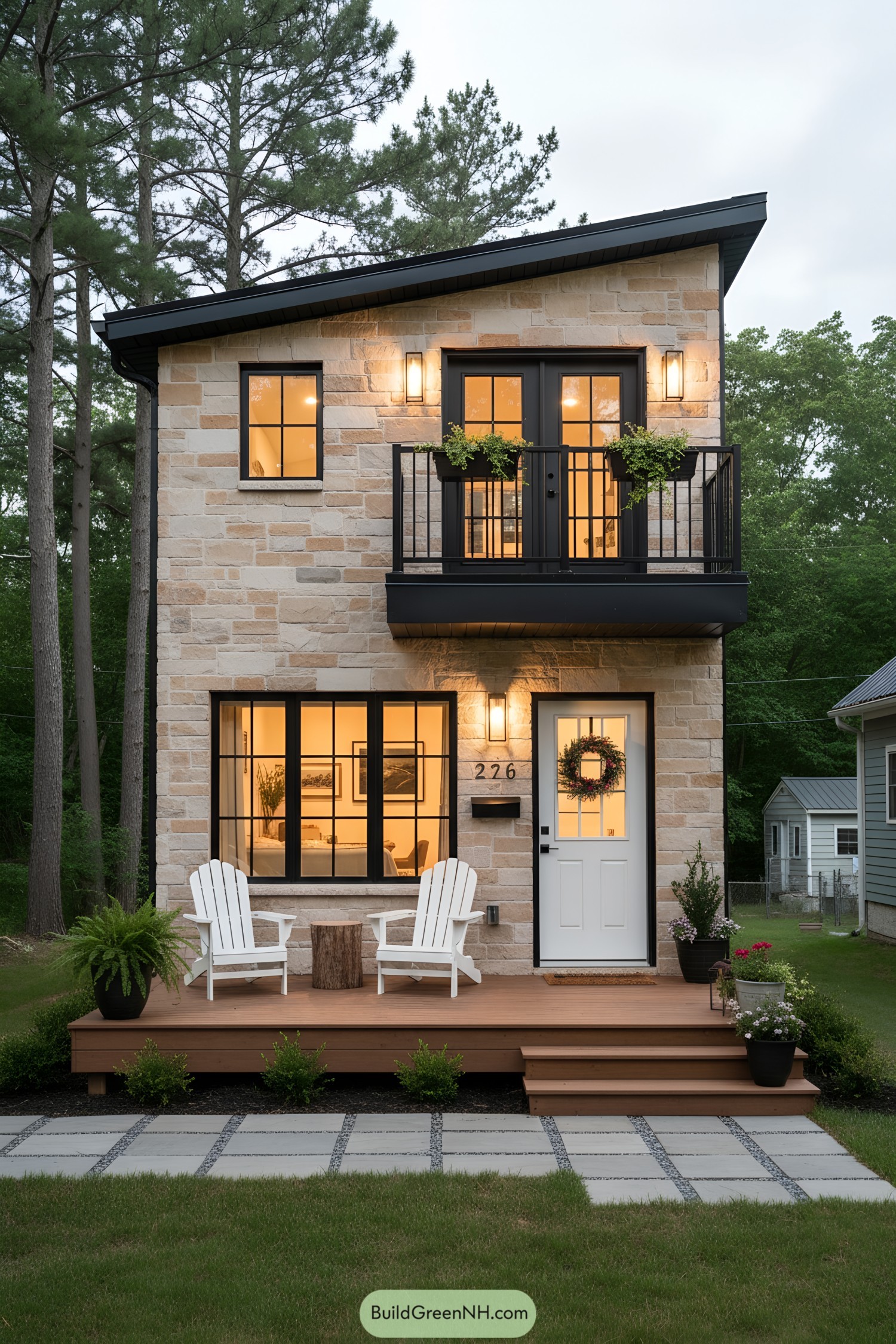 Two-story stone tiny house with black-framed windows, small balcony, and warm porch lighting