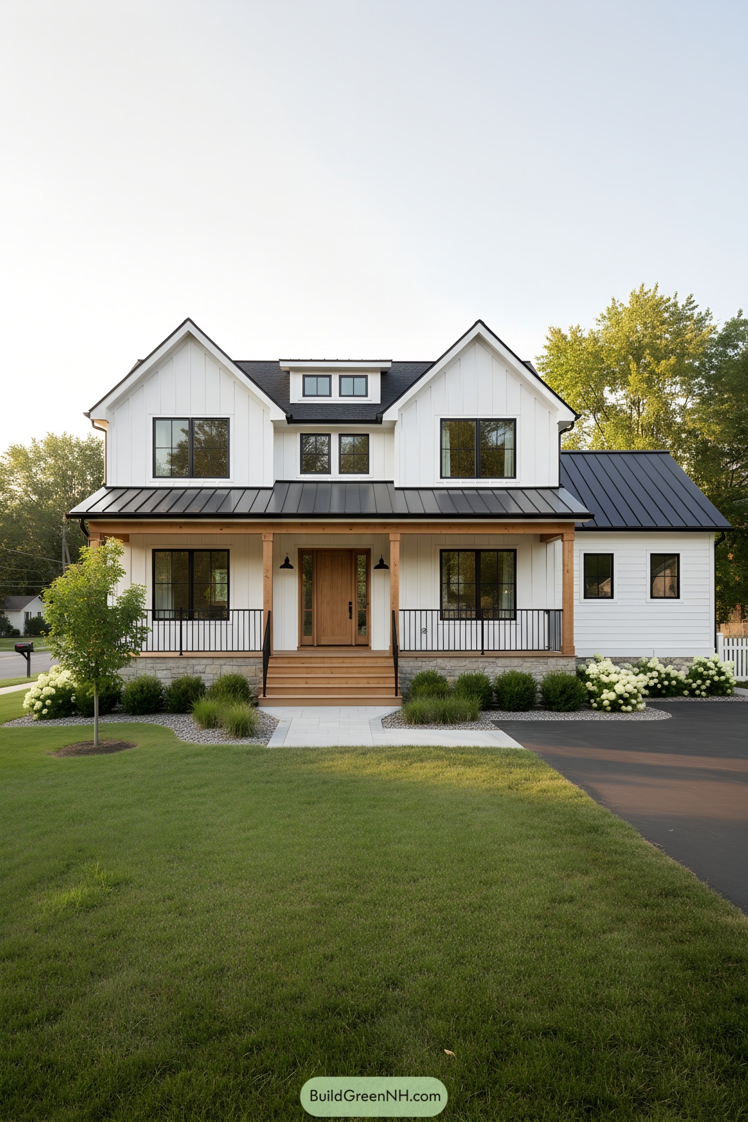 White farmhouse with black metal roof, gabled fronts, and a warm wood porch framed by simple landscaping