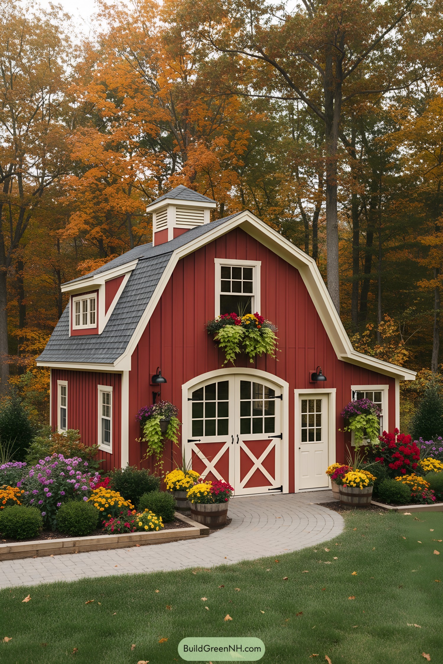 Red gambrel tiny barn with cupola, white trim, and flower-filled planters in a landscaped yard
