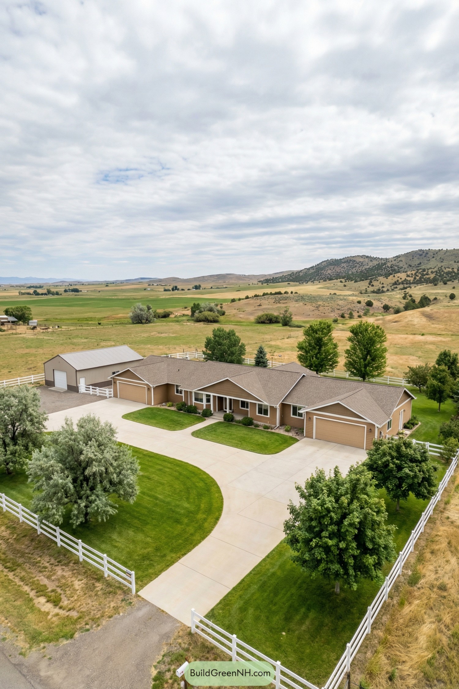 high-res aerial photo of rural house country living, long one-story ranch-style facade with multiple connected wings and an adjacent rectangular outbuilding, exterior in warm tan-brown siding with subtle texture and white trim, simple horizontal massing with low profile and modest projections, structure mainly rectangular with shallow offsets and attached garages at both ends, materials include wood or fiber-cement siding, concrete foundation, and light-colored concrete driveway and walkways, low-pitch gable roofs with brown asphalt shingles and minimal overhangs, mix of standard rectangular windows with white frames and grids plus a few larger picture windows facing the front yard, plain paneled garage doors in matching tan with minimal detailing, main entry door understated and sheltered by a small gable element, outdoor area features a broad curved concrete drive looping before the house and leading to garages and outbuilding, small planting beds with low shrubs and edging stones near the facade, expansive front lawn of vivid green grass framed by a white wooden ranch-style perimeter fence, scattered mature trees with full silvery-green and deep-green foliage around the house and along the drive, surrounding environment of open rural fields with patches of dry grass, gentle slopes, and low scrub-covered hills in the background, distant tree lines and hedgerows enclosing the property, wide sky with dramatic layered clouds creating soft diffused light, overall calm and picturesque countryside setting. real-life photo, high-resolution, architectural photography, soft lighting, cinematic composition.