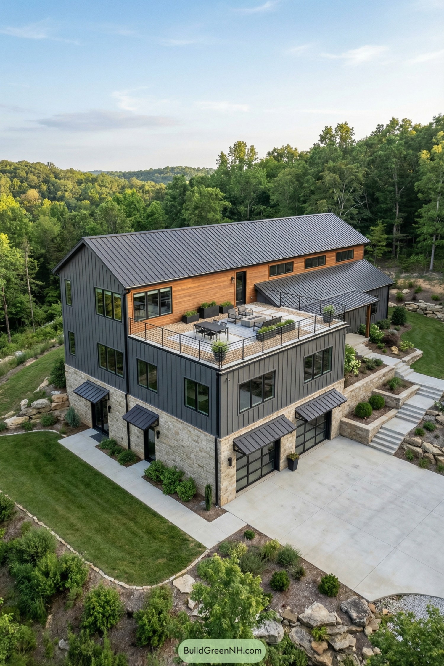 High-res aerial view photo of a modern rustic barndominium with rooftop deck; elongated L-shaped two-story structure above a partially exposed lower level, combining vertical charcoal-gray metal siding, warm horizontal cedar cladding accents, and rough-cut beige-gray stone base. Dark standing-seam metal gable roofs with moderate pitch, simple eaves, and minimal overhangs; secondary shed roofs over lower elements. Facade features large rectangular windows with dark aluminum frames, arranged in horizontal bands and corner groupings; some clerestory-style windows on upper level; black-framed glass garage doors on the stone lower level, each with its own shed metal awning. Main entrance defined by stone columns and a covered porch under the upper volume; additional side decks with simple black metal railings and wood plank flooring. Rooftop deck above the stone garage level, edged with slim black metal railing, surface divided into light pavers and gravel, furnished with black outdoor chairs and table, a few low planters, and a central seating zone. Outdoor areas include wide concrete driveway and parking pad, stepped concrete paths, retaining stone walls, and tiered terraces connecting levels. Landscaping with neatly edged lawn, low shrubs along foundations, natural rock outcrops, and minimal ornamental plantings, transitioning into dense green forest. Surrounding environment of rolling wooded hills and distant ridgelines under a bright blue sky with soft clouds, creating a secluded, picture-worthy mountain setting. real-life photo, high-resolution, architectural photography, soft lighting, cinematic composition.