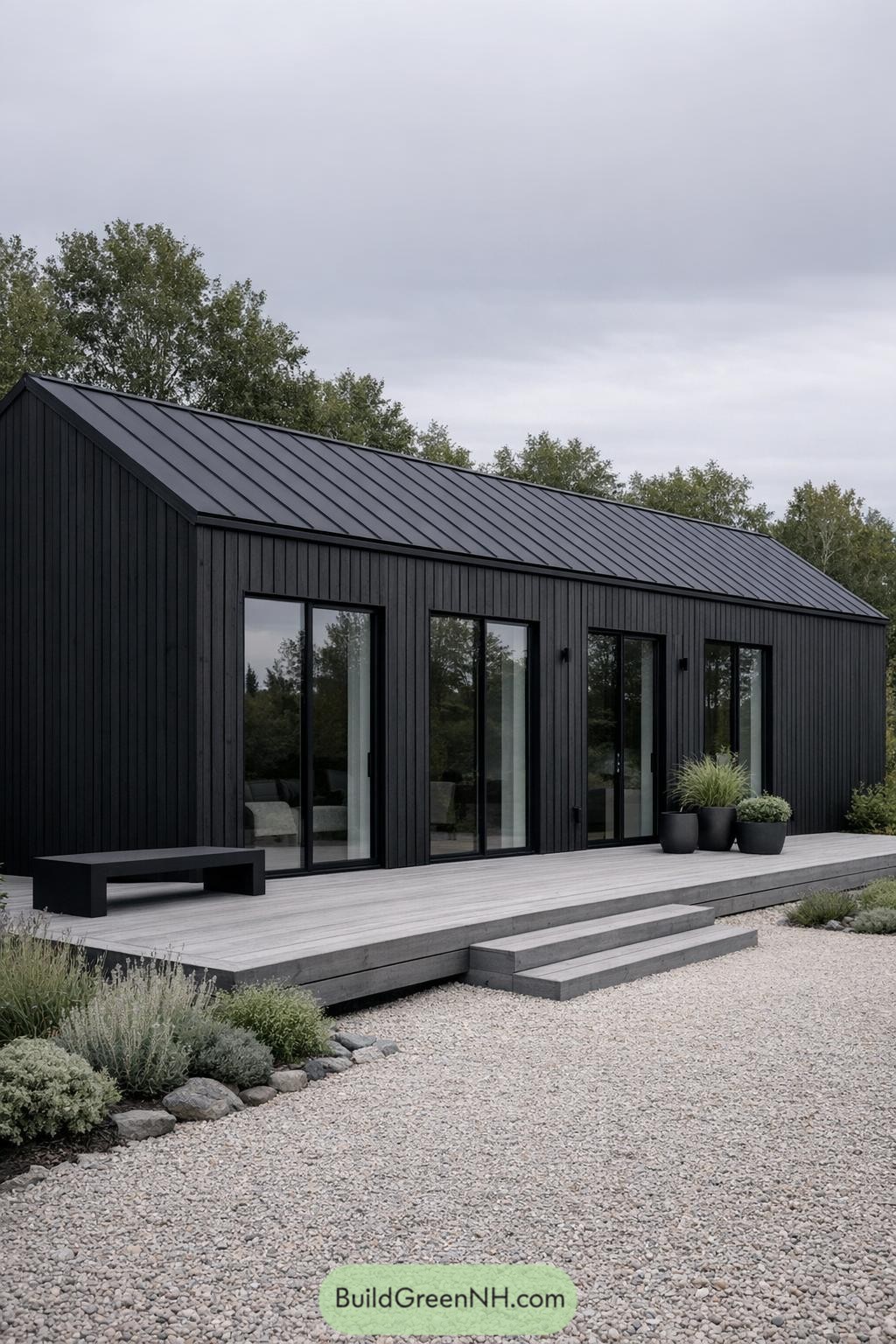 Black timber-clad Scandinavian cabin with metal gable roof, full-height glass doors, and a low gray deck opening to a gravel courtyard