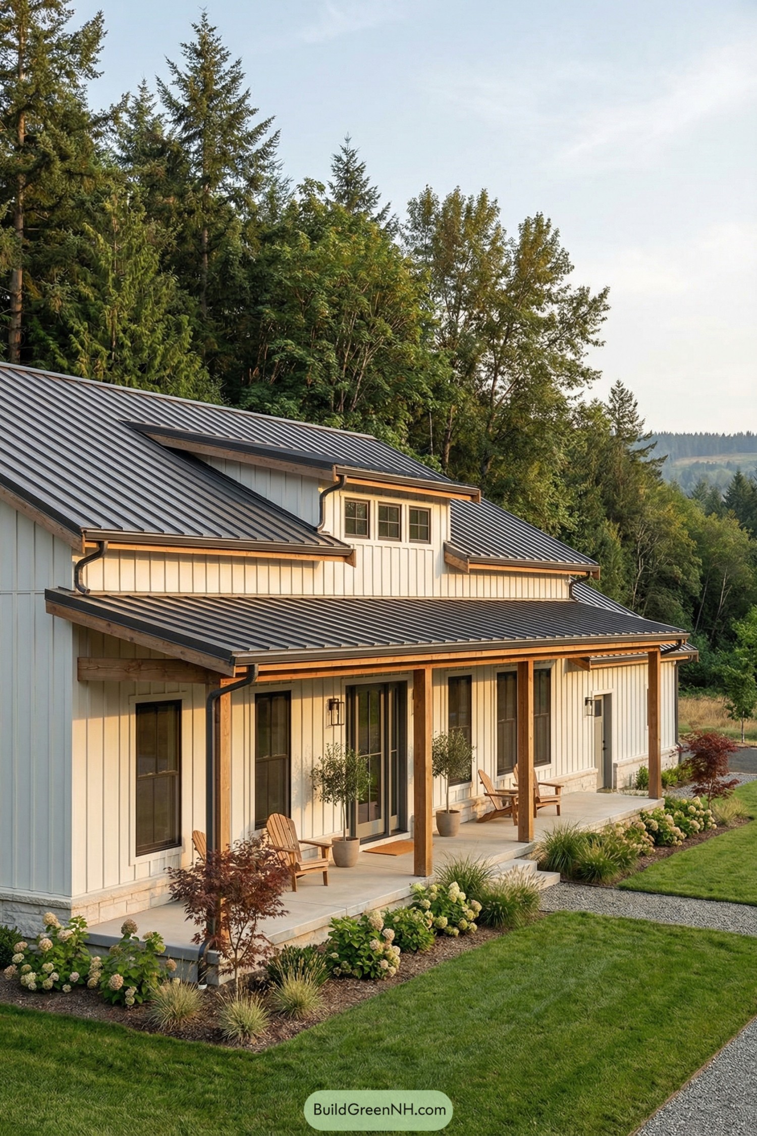 White barndominium with metal roof and long covered porch facing landscaped yard and forest backdrop