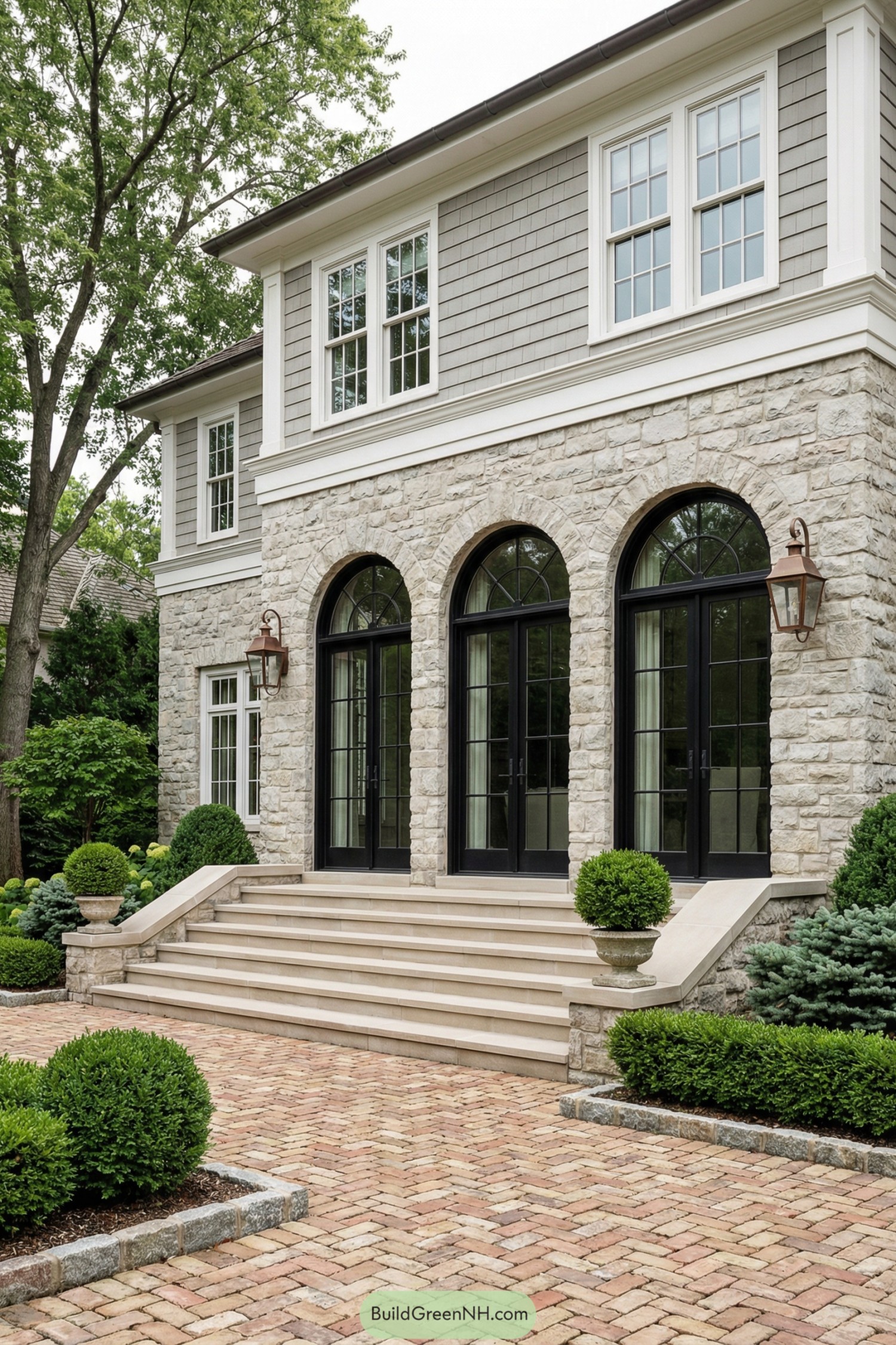 Stone facade home featuring three tall black arched doors, wide front steps, and manicured shrubs along a brick courtyard