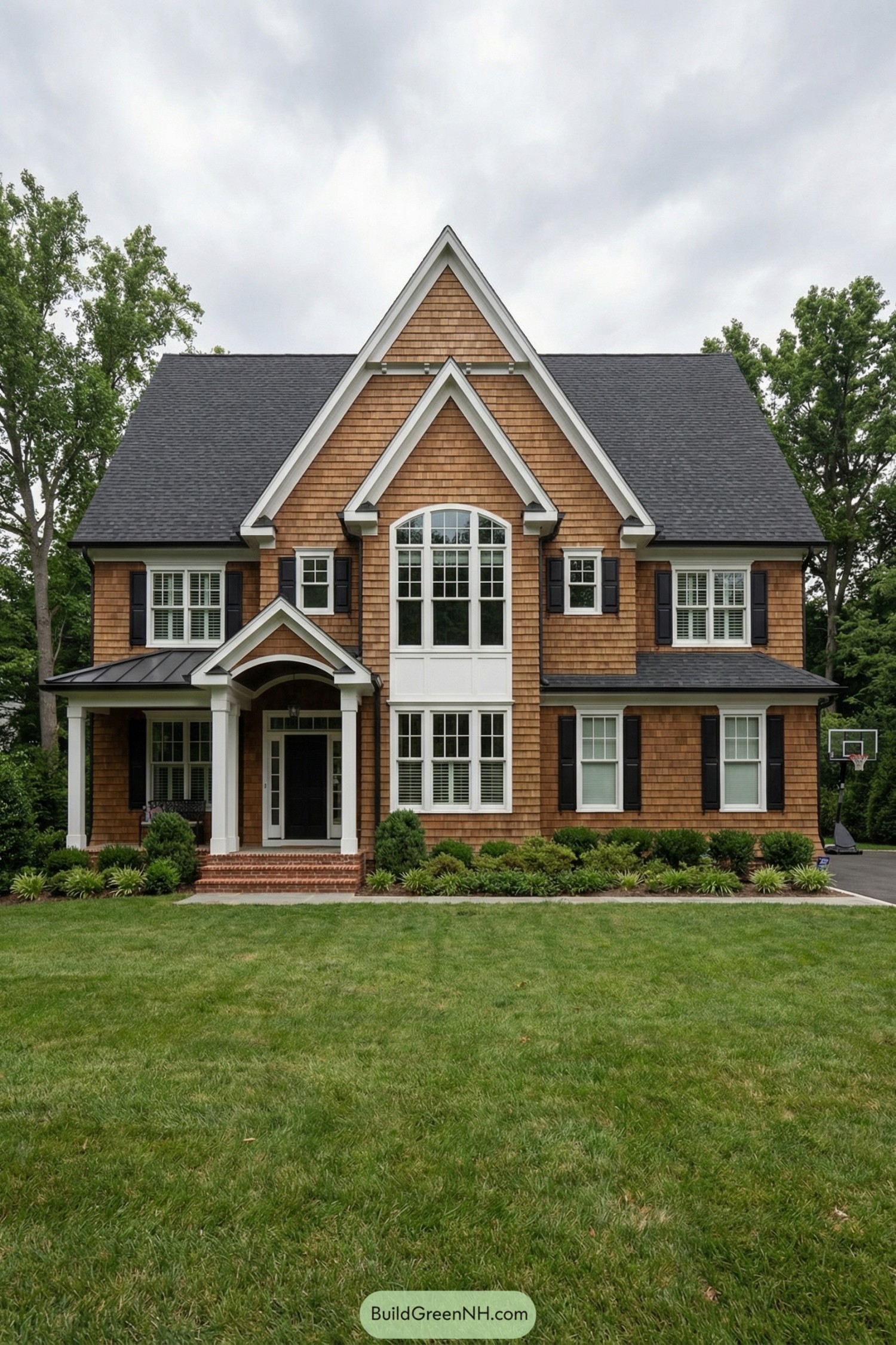 Two-story shingle-style house with steep gables, central arched window, and white-trimmed porch framed by neat foundation landscaping. Wide lawn in front with driveway and trees surrounding the home