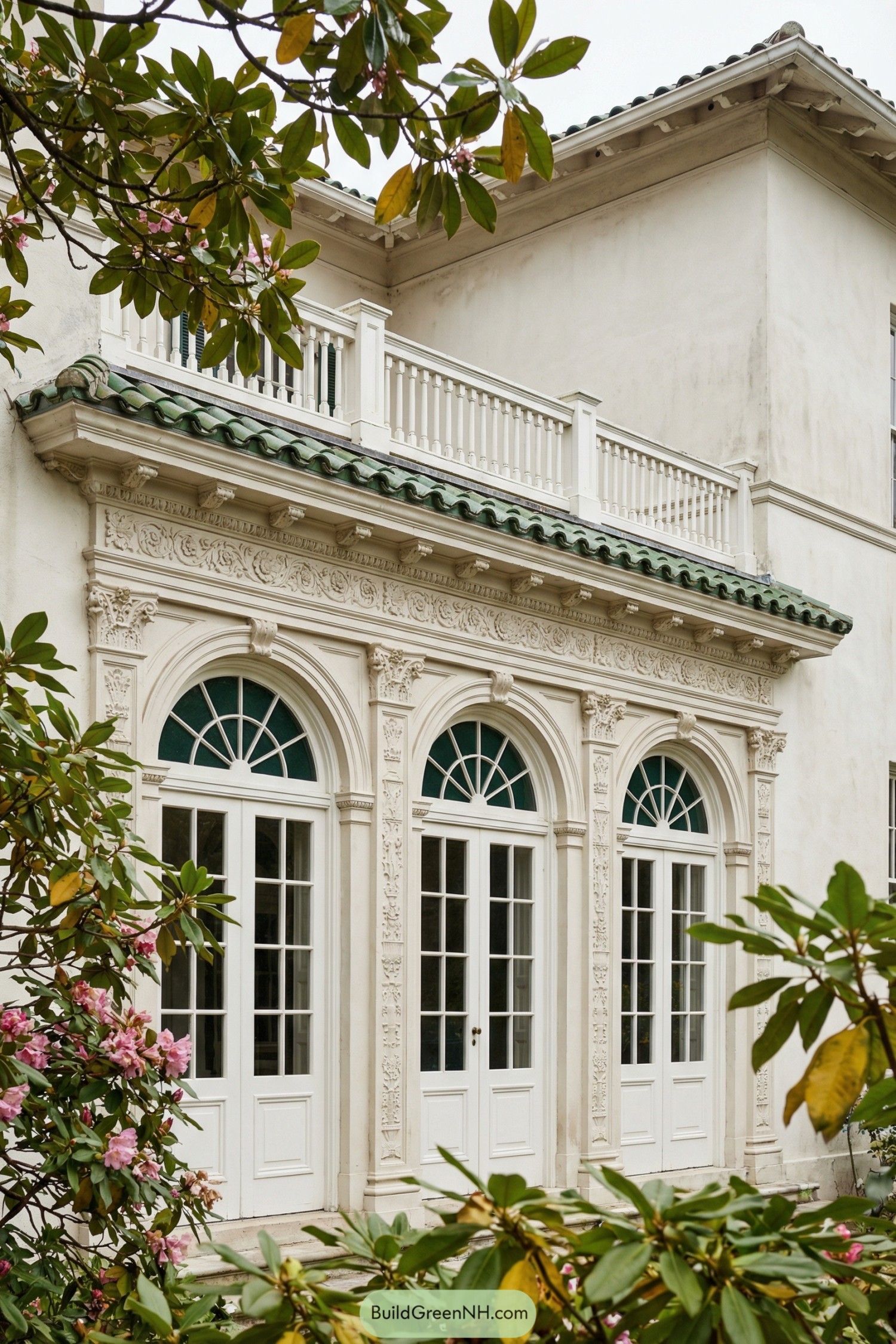 Ornate stucco facade with triple arched French doors and green tile roof above garden shrubs