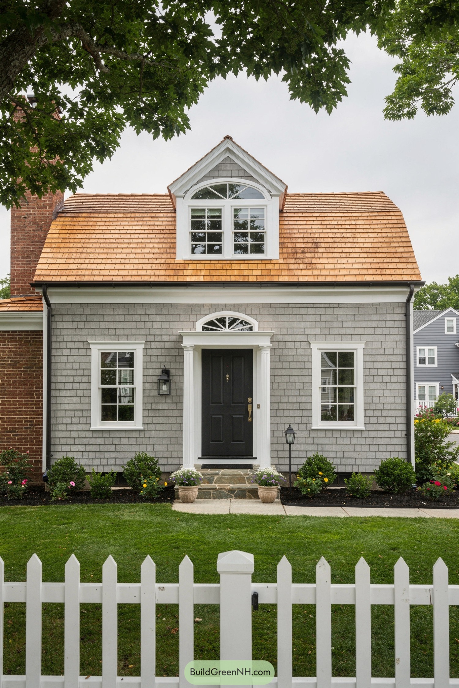 Small gray shingle house with cedar roof, dormer, and white picket fence