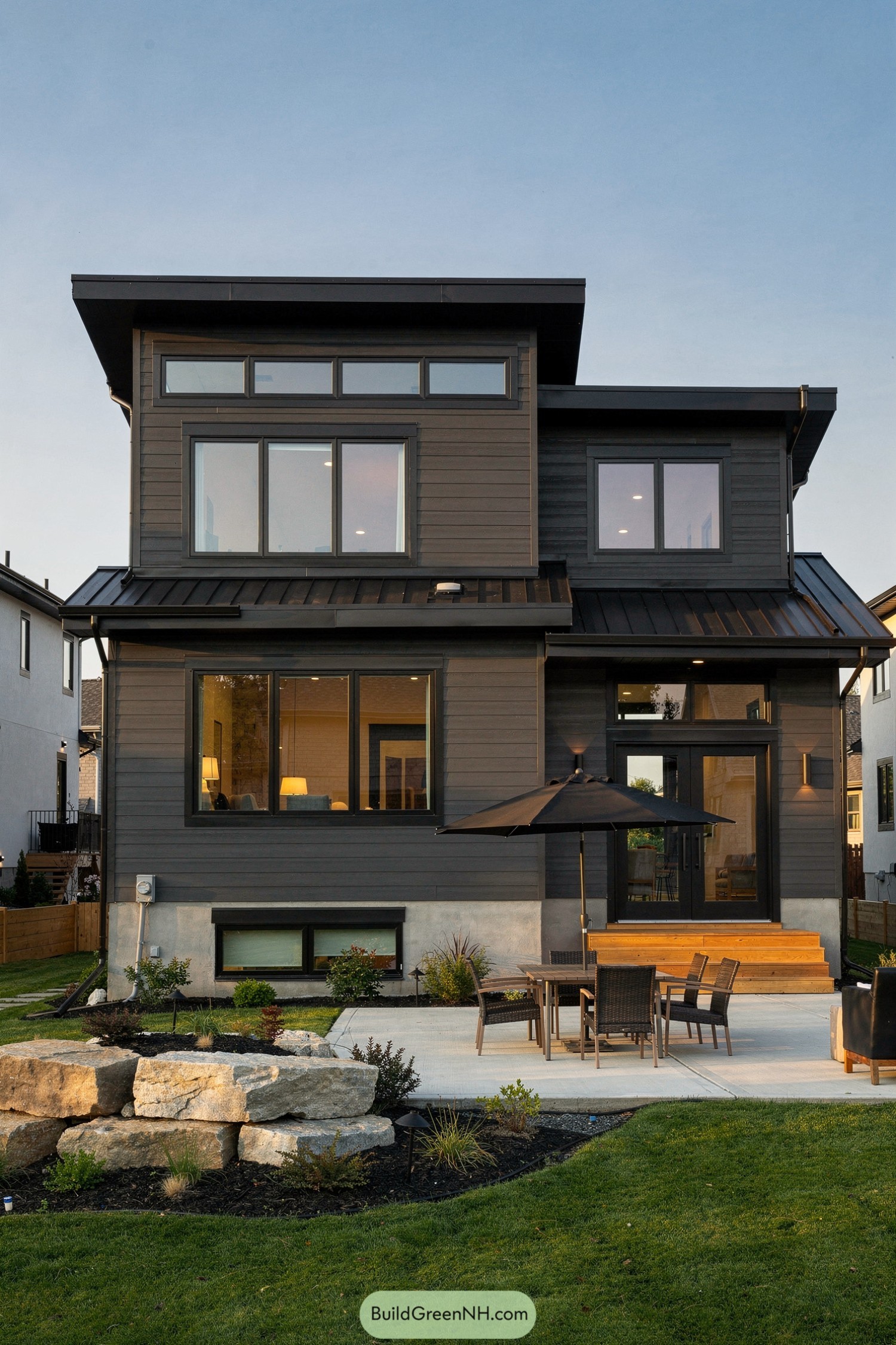 Dark charcoal siding wraps the stacked volumes of a modern two story home with wide windows and a clean lined shed roof. A concrete patio with outdoor dining set and umbrella extends into a neatly landscaped backyard with boulders and fresh lawn
