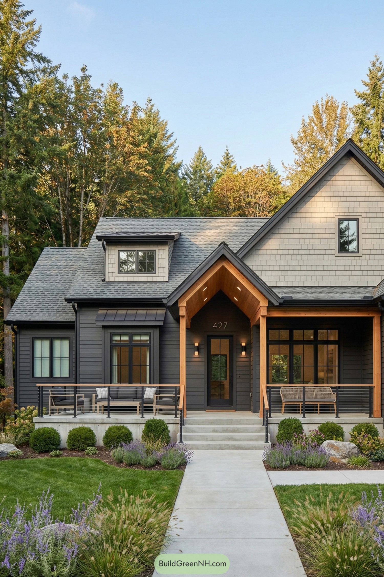 high-res photo of house with Porch Gable, modern farmhouse ranch facade with long front porch and prominent front-facing porch gable over the entry, dark charcoal horizontal lap siding on the main walls, light warm-gray shingle siding infill on the tall right gable and small central dormer, black trim and gutters, low and wide rectangular shape with a single main story and one steep front gable mass on the right, grey architectural asphalt shingle roofing with simple ridgelines and a small shed-roofed dormer window at center, large black-framed grille windows including paired casements on the left wing, a central box bay window with a small dark metal shed roof, a wide multi-pane picture window on the right porch area, and a compact window in the upper right gable, single dark entry door beneath the porch gable with a vertical glass panel, side-mounted modern wall sconces, and minimalist address numbers above, deep front porch spanning center and right sections with warm natural-wood posts and beams, wood soffit under the porch gable, recessed ceiling lights, slim horizontal cable or metal railings with wood top rails, concrete porch floor and broad concrete steps leading up from the front walk, outdoor seating group with sofa and chairs on the left side of the porch and woven bench seating on the right, layered modern landscaping with manicured lawn, low rounded boxwood shrubs, ornamental grasses, lavender and purple flowering perennials, accent boulders, and structured planting beds along the foundation and at the sidewalk edge, straight concrete walkway at the front bordered by mixed grasses, backdrop of dense tall trees with a mix of evergreens and deciduous foliage in greens and muted autumn tones under a clear blue sky, overall clean and picturesque suburban woodland setting, real-life photo, high-resolution, architectural photography, soft lighting, cinematic composition.