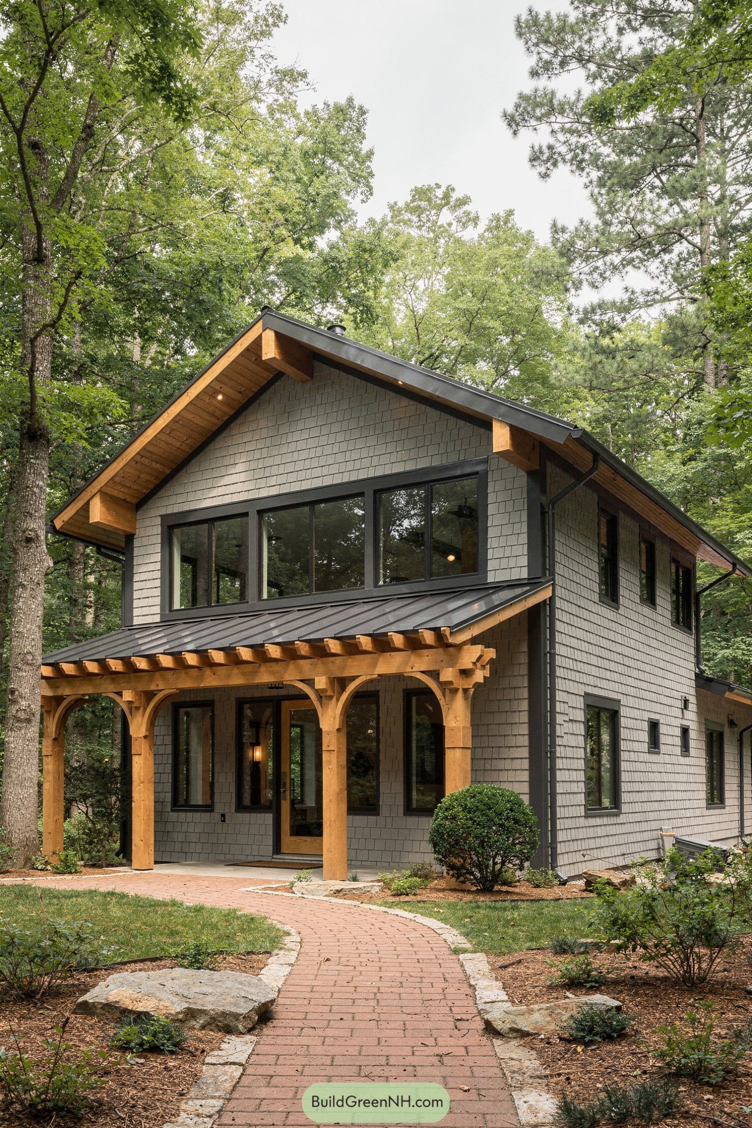 high-res photo of house with Shingle Siding, two-story rectangular cottage-style facade with modern lines, light gray irregular shingle siding on all walls, dark charcoal trim and corner boards, compact gable volume with a slightly lower flat-fronted section along the main facade, materials include painted wood shingles, smooth wood trim, and exposed natural-stain timber brackets and beams, dark charcoal standing-seam metal gable roof with shallow pitch and small shed-style metal roof above the main entry, continuous band of tall horizontal casement or fixed windows across the upper front level with dark frames, additional single and paired rectangular windows on sides and rear with matching trim, narrow glass-panel entry doors with dark frames at front and side, extended wood pergola structure running the width of the front facade with heavy timber posts and curved brackets, brick-paver walkway and small entry stoop leading to the door, low natural stone edging and scattered flat stones near the path, informal landscaping with small shrubs, groundcover, and a clipped round bush near the side door, house set in a densely wooded environment with tall mature trees and layered green foliage surrounding and rising behind the building, soft overcast daylight creating a calm, picture-worthy forest setting. real-life photo, high-resolution, architectural photography, soft lighting, cinematic composition.