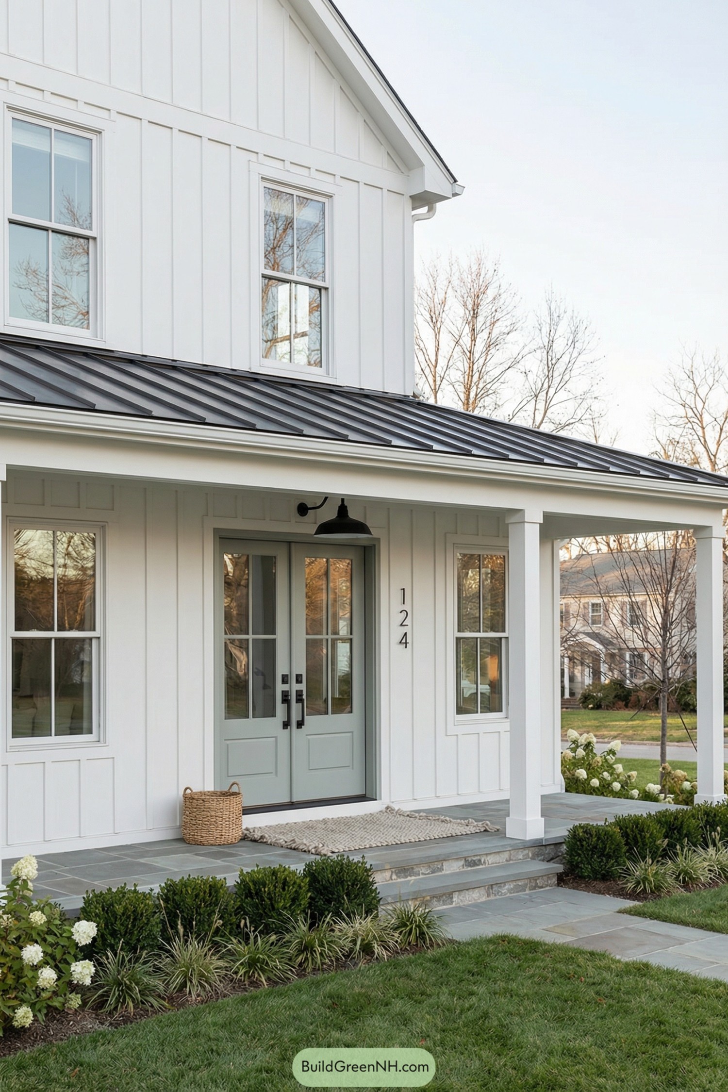 high-res photo of house with Shiplap Siding, two-story modern farmhouse facade with clean lines and a deep covered front porch, vertical white shiplap cladding on all exterior walls with crisp white trim and simple corner boards, soft sage-gray centered double entry doors with upper nine-lite glass panels and recessed lower panels, matte black rectangular door hardware, thin black metal house numbers mounted on the siding beside the doors, large black metal gooseneck porch light above, medium-pitch front gable roof with dark charcoal standing-seam metal and plain white fascia, evenly spaced double-hung windows with white frames and divided lites on both floors flanking the entry, wide stone steps leading to a gray slate-tiled porch floor, simple square white porch columns, woven natural rattan basket and chunky knit neutral outdoor rug near the threshold, low foundation planting of boxwood hedges, ornamental grasses, and white hydrangeas along the base, neat green lawn extending to the foreground, scattered leafless deciduous trees and a distant traditional house softly blurred in the background, clear sky and soft daylight giving mild reflections in the glass. real-life photo, high-resolution, architectural photography, soft lighting, cinematic composition.