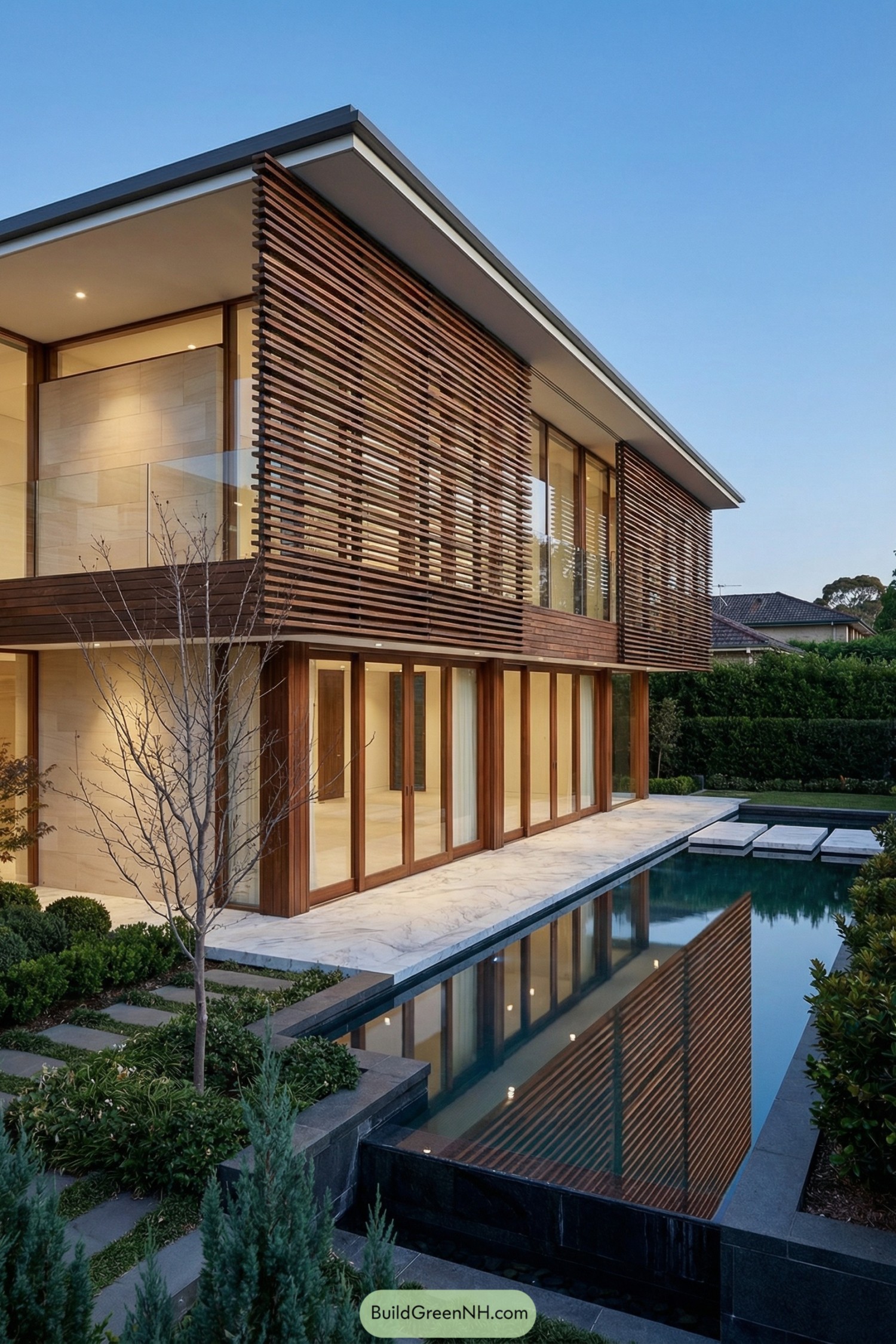 high-res photo of house with Timber Slats, two-story modernist facade with strong horizontal lines, upper floor wrapped in dense irregular horizontal timber slats forming a continuous screen, slats in rich warm walnut tone with varied spacing, facade behind slats composed of large floor-to-ceiling glazing and smooth light stone panels, colors dominated by warm wood, light beige stone, clear glass, dark water reflections, building mass rectangular with clean sharp edges and deep cantilevered terraces, materials including natural timber, honed white-veined marble for platform and terrace edges, smooth light stucco or stone side walls, dark stone cladding at water level, flat roof with thin projecting eaves and crisp white underside, expansive sliding windows and glass walls framed in dark wood, some panes clear and others lightly frosted, tall timber-framed sliding glass doors aligned in a row on ground floor opening onto marble terrace, outdoor area featuring a narrow reflective lap pool hugging the facade on two sides with mirror-like still water, marble terrace slabs floating above water with precise joints, low glass balustrades near corners, landscaping with manicured groundcover, low sculptural shrubs and small conifers along pool edge, a slender leafless tree and layered planting beds with stepping stones further out, surrounding background of dense green hedges, mature trees, and distant residential roofs under clear blue evening sky, overall composition calm, symmetrical, and highly picture-worthy with reflections of timber slats and sky in the water. real-life photo, high-resolution, architectural photography, soft lighting, cinematic composition