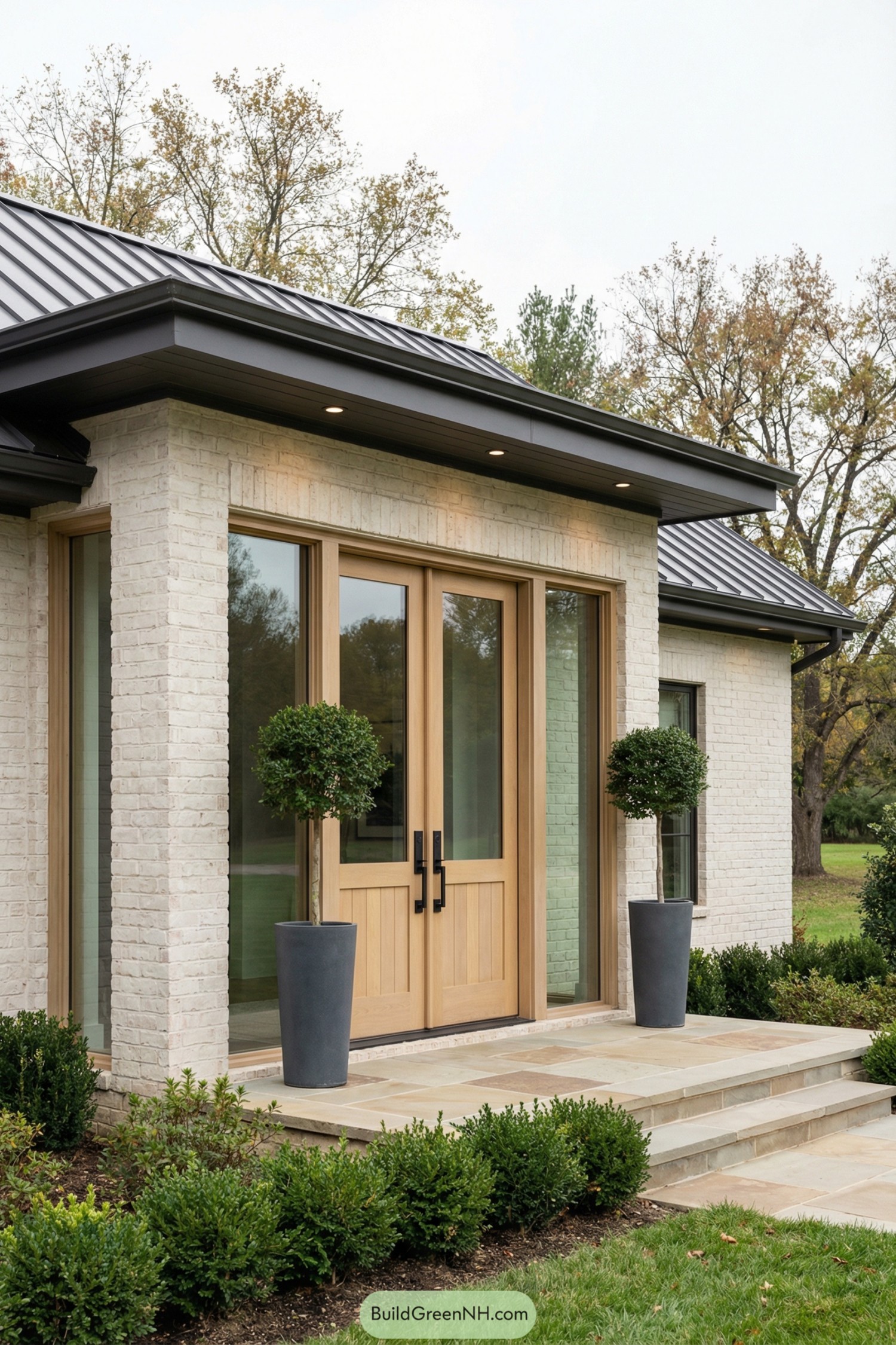 Light brick house front with pale wood double doors, tall sidelights, and two potted topiary trees on a stone stoop