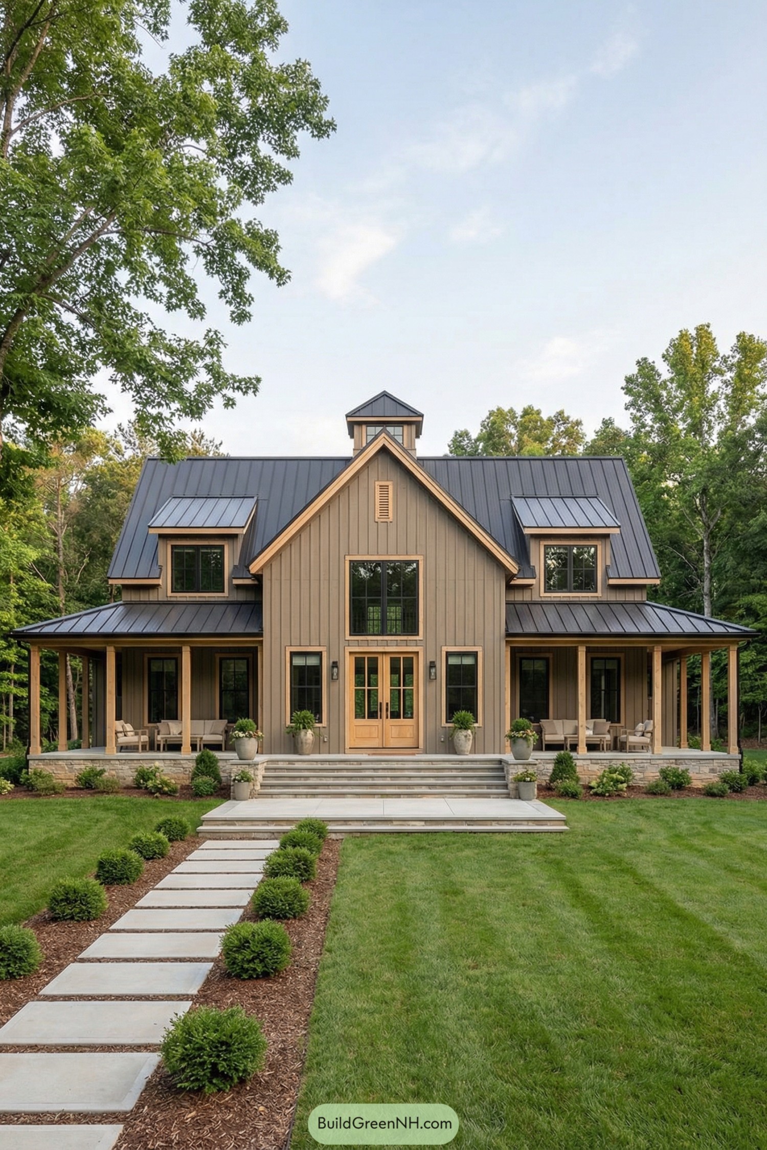 Two-story barndominium with wraparound porch and metal roof set in a wooded yard. Center path leads to wide steps, double doors, tall windows, and symmetrical dormers