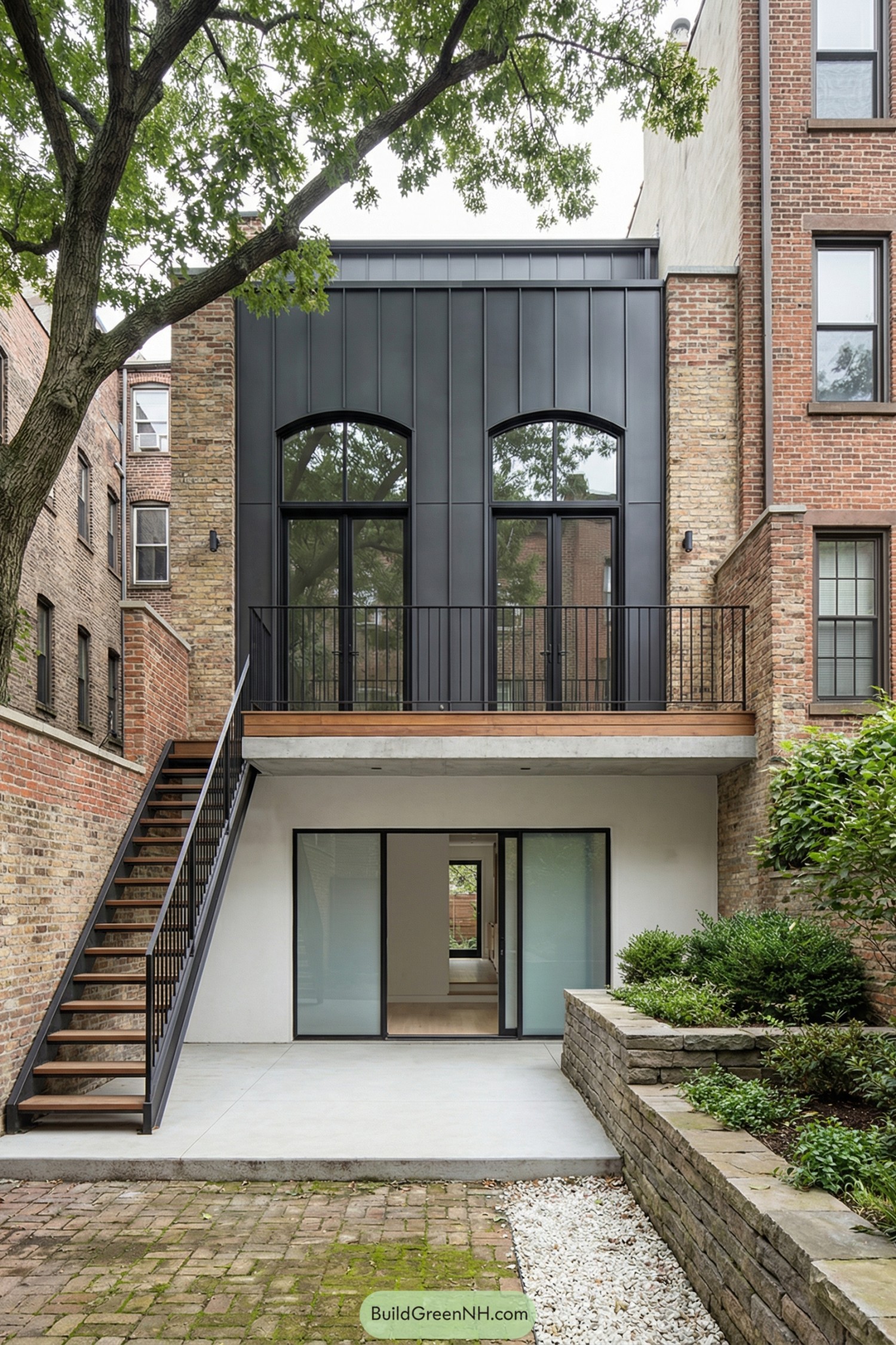 Rear courtyard view of a renovated brownstone featuring a dark metal-clad addition with tall arched windows, balcony, and garden