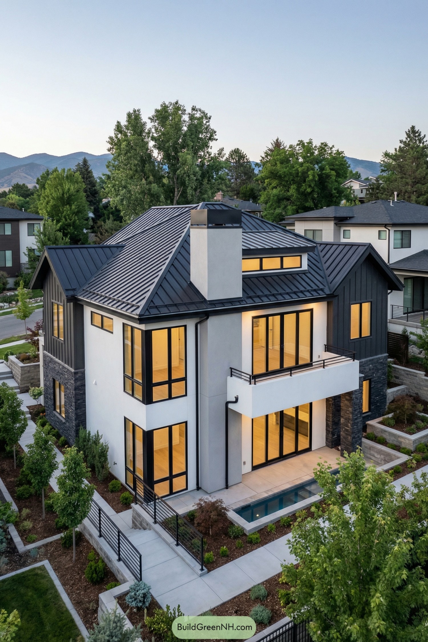 high-res photo of modern house with Black Metal Roof, facade, materials, colors, windows style, roof style, door style, outdoor area, landscaping, surrounding background. Two-story contemporary house, asymmetrical volumes, clean lines, slightly elevated on a sloped lot. Facade combines white smooth stucco on lower level with dark charcoal vertical siding and dark stone accents on corners and base. Exterior color palette: black, charcoal gray, crisp white, light natural wood at soffits and balcony ceilings. Complex gabled structure with multiple intersecting roofs, all standing-seam black metal panels, medium pitch, sharp eaves, minimal overhangs, black gutters and downspouts. Central light-gray rectangular chimney with black metal cap rises above main ridge. Large floor-to-ceiling windows in black aluminum frames, mix of fixed and sliding panels; corner glazing and wide horizontal strip windows on upper level. Black-framed glass doors: full-height sliding doors opening to balcony and patio, with narrow sidelights, very minimal trim. Balcony with white solid parapet and slim black horizontal metal railing, projecting from upper floor. Lower level with small rectangular reflecting pool or plunge pool integrated into terrace edge, framed by white walls. Paved walkways in light gray concrete stepping down the slope, black metal railings along stairs and edges. Landscaping with structured yet lush look: neatly pruned small trees, ornamental shrubs, linear planting beds with mulch, low groundcover, manicured lawn patches; stone retaining walls and raised planters matching house stone. Surrounding environment is a green residential hillside with tall mature trees, neighboring modern houses partially visible, distant low mountains under soft evening light, clear sky. real-life photo, high-resolution, architectural photography, soft lighting, cinematic composition