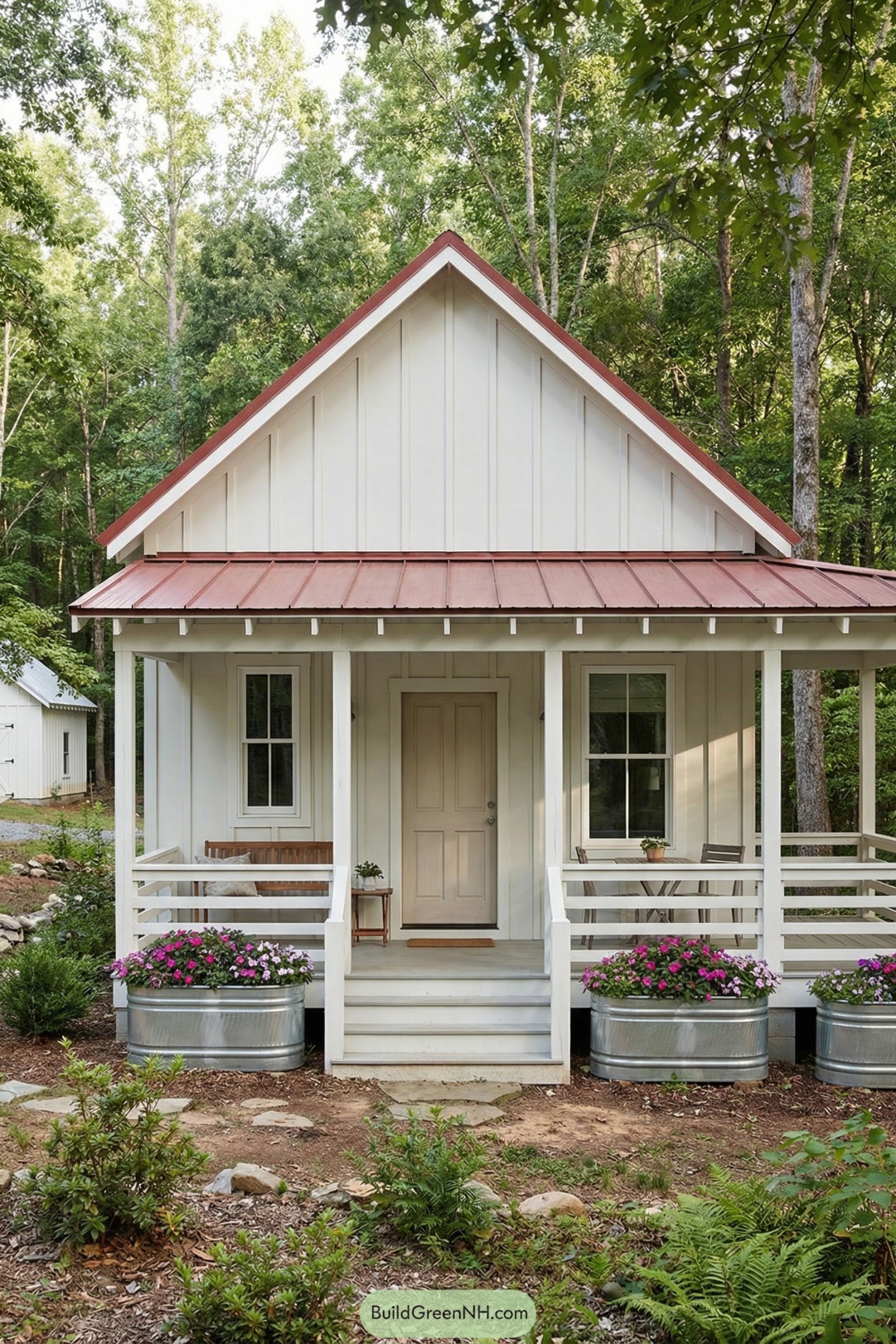 Small white board and batten cottage with red metal roof and flower-lined front porch in a wooded clearing