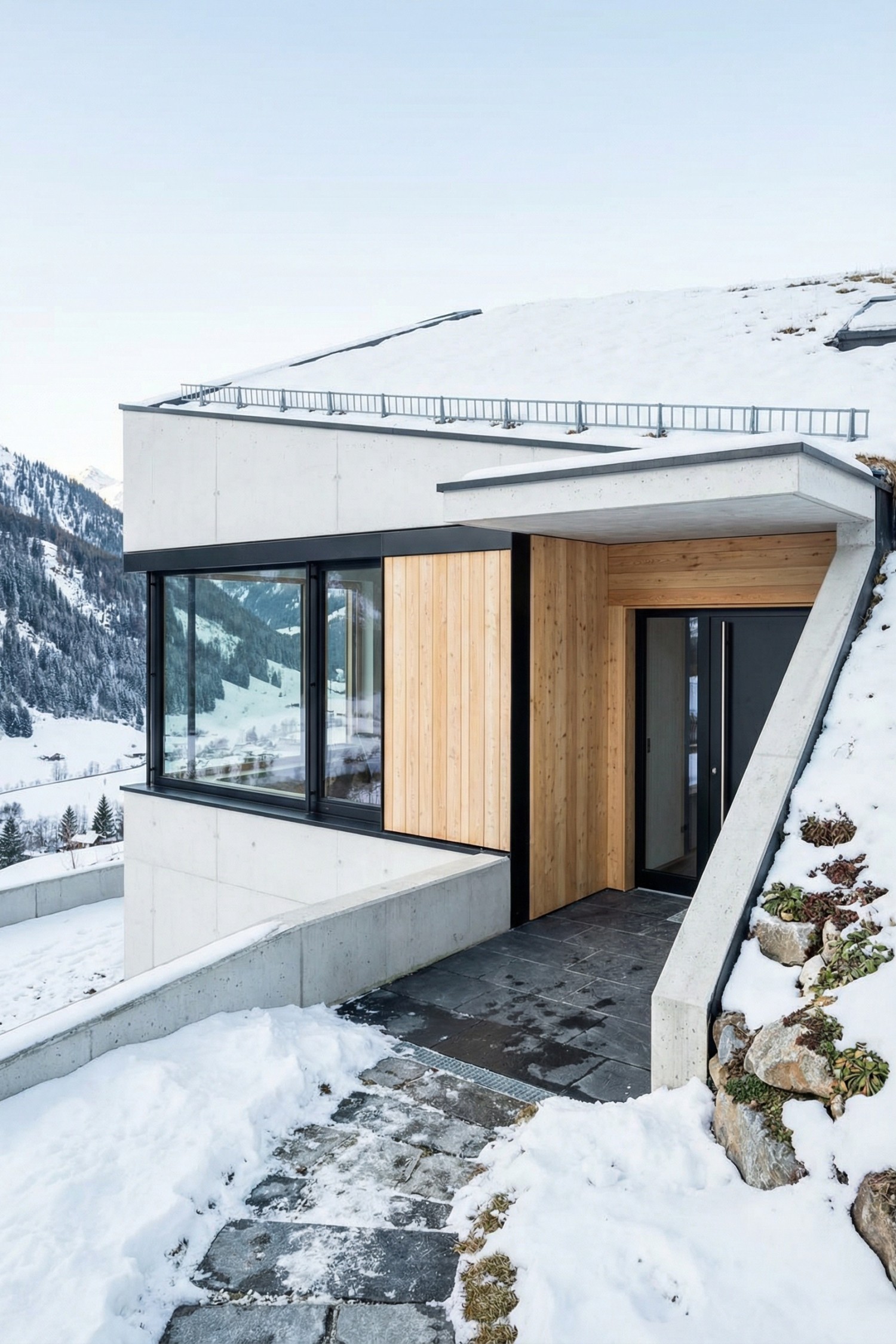 Concrete and wood-clad earth sheltered house set into a snowy hillside with large corner windows