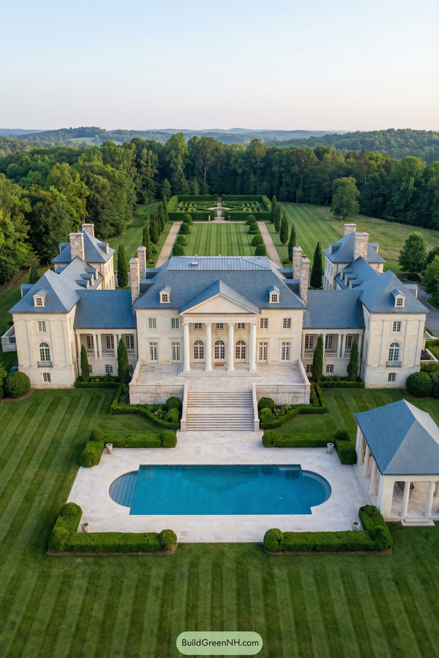 high-res aerial view photo of Classic Mansion, grand symmetrical neoclassical château-style facade in light cream stone or stucco with a central two-story portico of tall white columns and triangular pediment, long lateral wings with slightly projecting end pavilions, smooth walls with minimal ornament, subtle cornices and balustraded terraces, exterior colors dominated by cream walls, white trim, and slate-blue roofing with pale stone paving, elongated rectangular building footprint with shallow courtyards and a central garden axis leading to a formal pool, steep mansard and hipped slate-blue roofs with multiple ridges, dormers with pediments, flat rooftop sections with skylights and discrete chimneys in matching light stone, tall French windows arranged in strict symmetry with white frames, some arched on the ground level and rectangular above, numerous full-height French doors opening to terraces and garden, central grand entrance doors recessed behind the columned portico, additional arcaded loggias and columned pavilions at the sides, outdoor area with broad central stone staircase descending from the main terrace to a formal rectangular swimming pool with a rounded end, pale stone pool deck and flanking clipped hedges, secondary paths and platforms in light stone connecting to a detached pavilion with open colonnade and matching roof, meticulous landscaping with wide striped lawns, sculpted box hedges, spherical and conical topiary, tall cypress-like trees, large evergreen conifers, and a formal geometric hedge garden in the distance, surrounding environment of dense forest edge enclosing the estate and soft rolling hills on the horizon under clear calm sky, overall composition centered on the mansion and axial garden layout, single real-life photo, high-resolution, architectural photography, soft lighting, cinematic composition, strictly no collages.