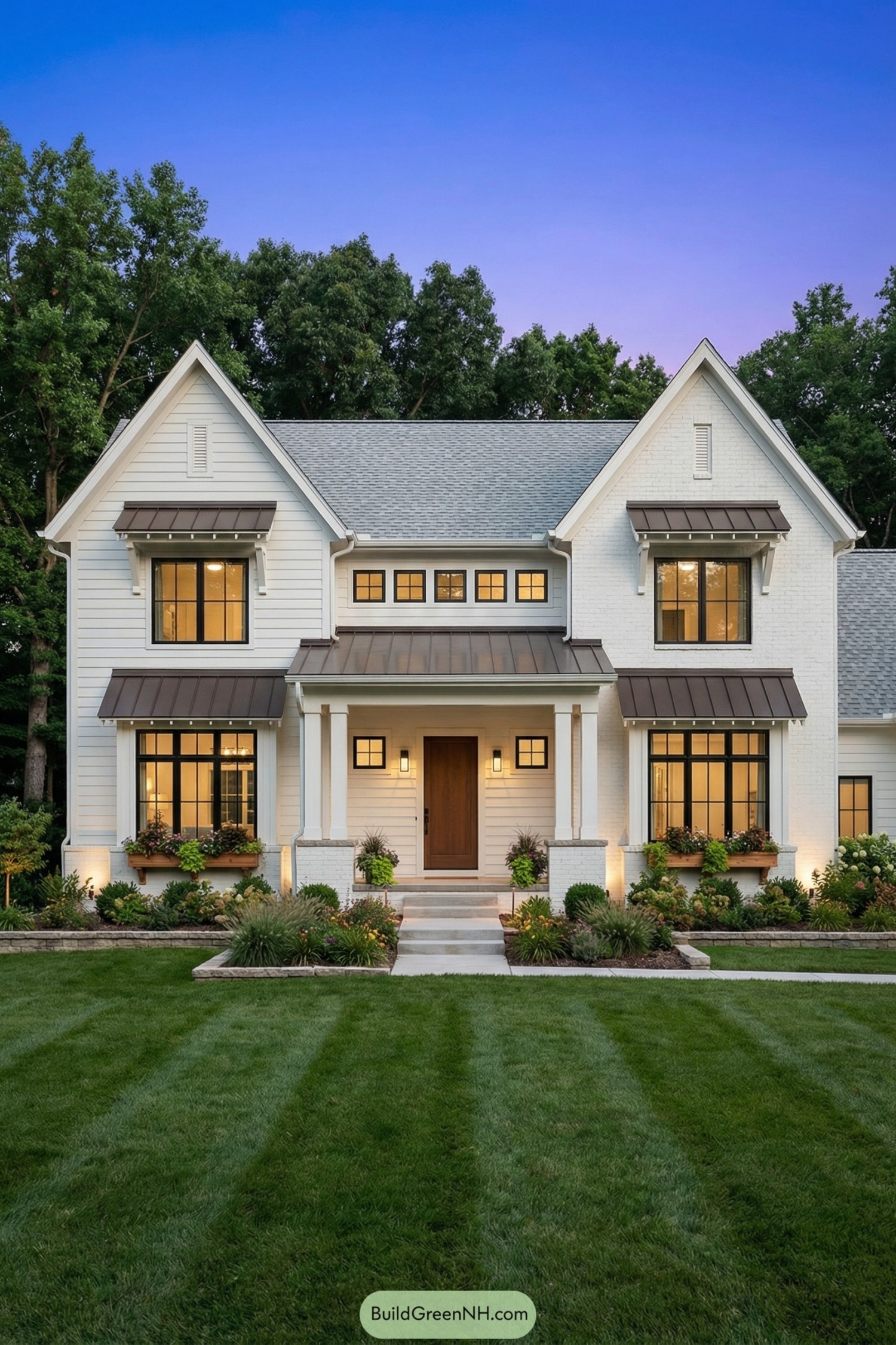 Two story white modern farmhouse with gray roof and bronze metal awnings. Symmetrical front elevation with warm lit windows and lush landscaped yard