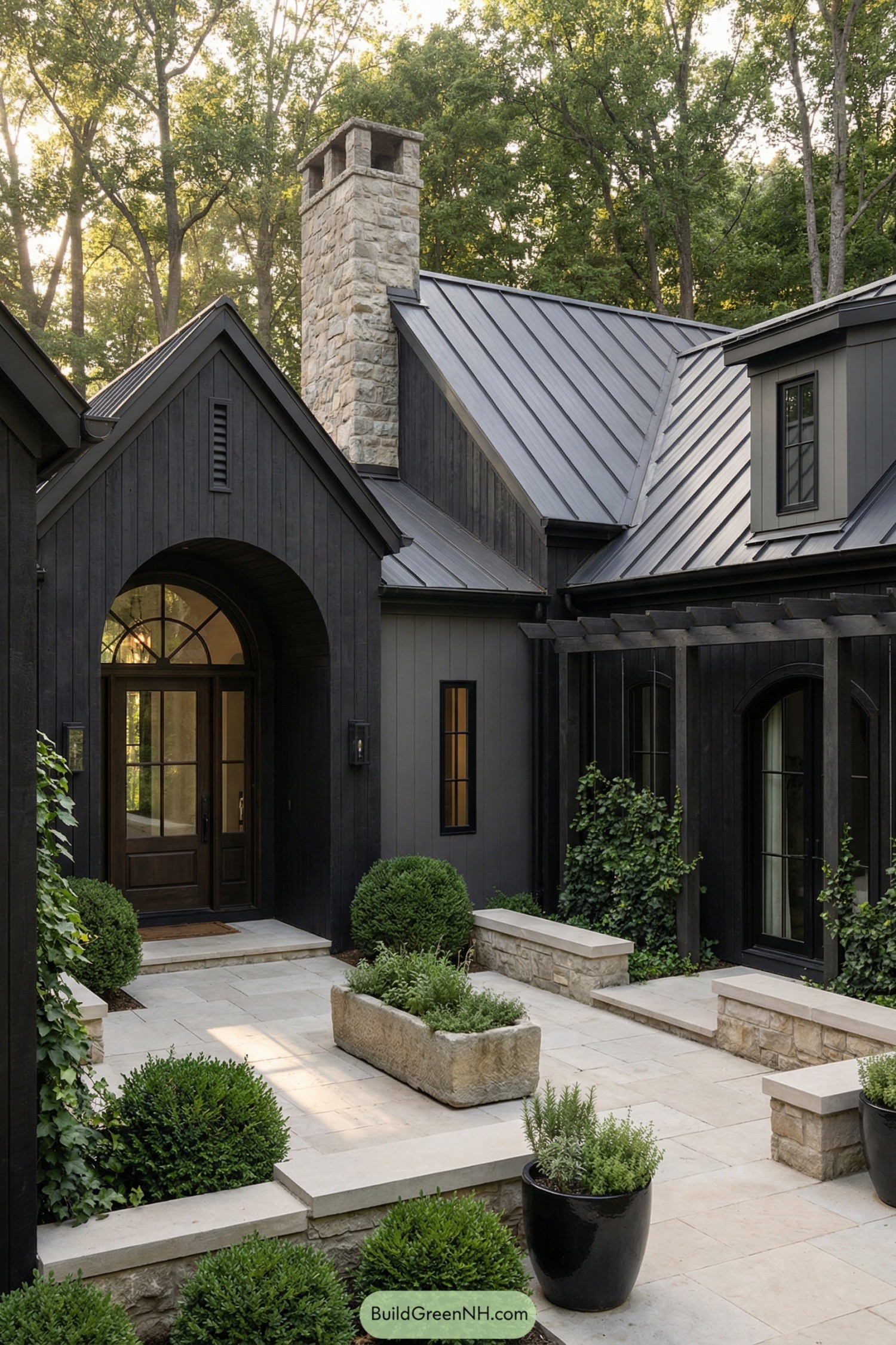 Dark modern cottage with black siding, metal roof, stone chimney, and lush courtyard plantings