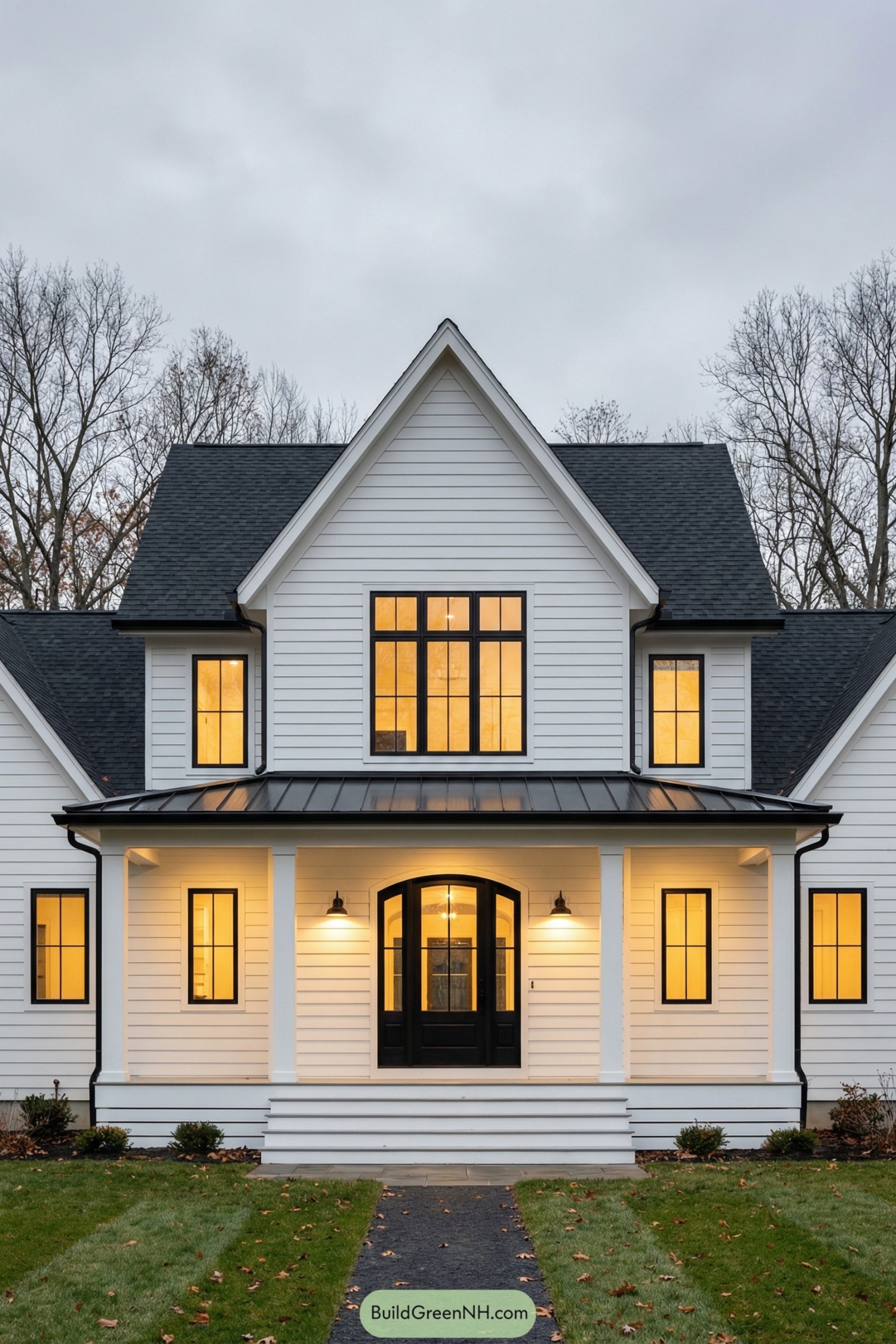 high-res photo of black and white house exterior, modern farmhouse facade with clean symmetrical front and central front-facing gable, crisp white horizontal siding, strong black trim and window frames, tall rectangular massing with a main two-story volume and lower side wings, simple rectangular footprint with deep front porch running across the center. colors: white exterior cladding, black roof shingles, black framed windows and doors, black metal porch roof band, warm golden interior lighting visible through glazing. shape of the structure: two-story main block with steep front gable, lower side extensions with matching roof pitch, broad front porch platform raised slightly above ground with wide steps, simple rectilinear forms and sharp roof lines. materials of the building: painted wood or fiber-cement lap siding in white, smooth white corner boards and trim, black-painted metal window and door frames, black standing-seam metal accent roof over porch, asphalt shingle main roofing, white-painted wood porch columns and rail-free porch edge. roofing: steep gable roof in dark charcoal or black shingles, clean eaves with minimal overhangs, single main front gable centered on facade, side roofs receding with same pitch, subtle metal drip edges, flat black standing-seam metal strip above first-floor windows and porch. windows: tall, narrow, black-framed casement or fixed windows with multiple panes, evenly spaced in vertical alignment, three windows on second floor of main gable, paired groups on side wings, large grid-pattern windows flanking the entry on the first floor, warm reflections and interior chandeliers visible, no shutters. doors: central black front door with gentle arched top and large glass panel, matching black sidelights with divided panes, simple black hardware, full-height black-framed glass doors or sliders on either side of entry leading to porch. outdoor structures: wide white front porch with smooth decking and simple fascia, broad central steps leading from lawn to porch, white square columns supporting shallow porch roof, recessed downlights in porch ceiling, minimal black wall sconces on each side of front door, a few simple planters and lanterns arranged symmetrically near entry. landscaping: flat, well-kept lawn in front, thin dark mulch bed along porch edge, low ornamental grasses and compact shrubs near corners, a narrow dark stone or concrete path leading toward steps, scattered fallen leaves on grass for seasonal touch. surrounding environment: leafless tall trees framing the house in background, soft gray overcast sky, subtle depth of neighboring woodland suggested beyond, overall quiet suburban or semi-rural setting with no visible neighboring structures. setting and scene: straight-on eye-level view emphasizing symmetry and clean lines, early evening or late afternoon with warm interior glow against cool exterior light, calm and slightly moody atmosphere highlighting contrast between white facade and black elements. single real-life photo, high-resolution, architectural photography, soft lighting, cinematic composition, strictly no collages.