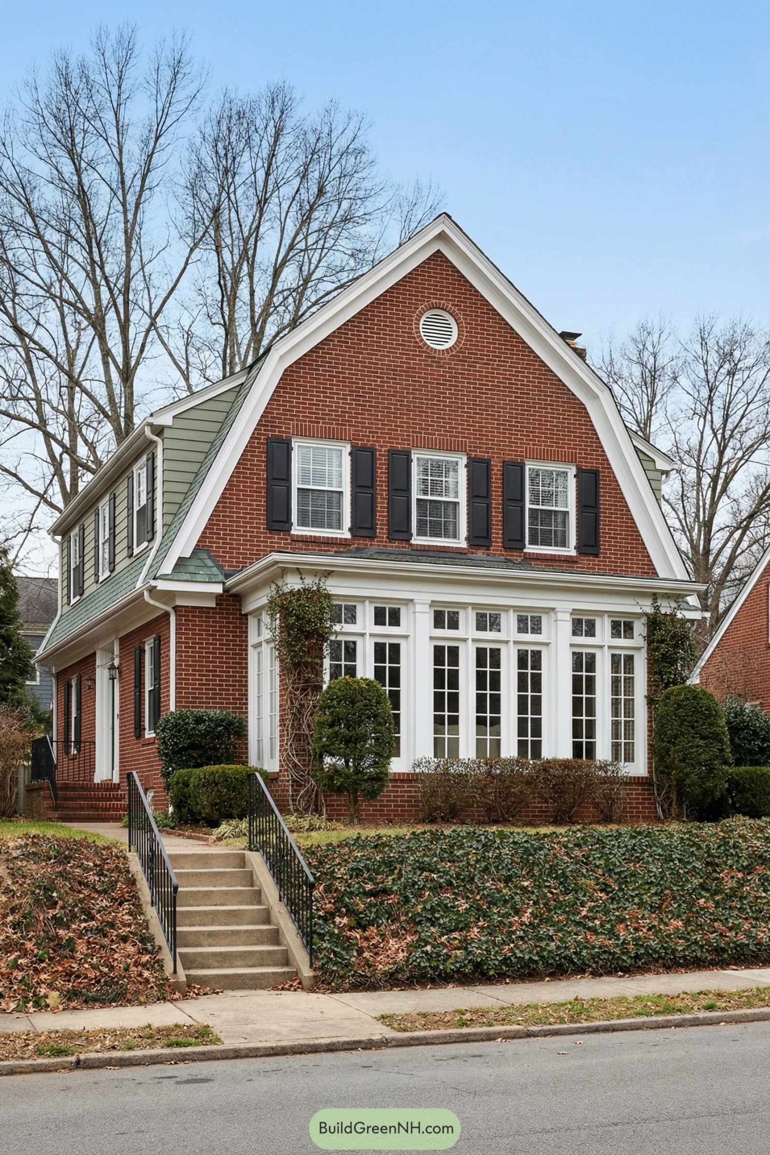 high-res photo of Brick Colonial House, symmetrical front facade of deep red brick with white trim, steep front-facing gable with clean white rake boards and a small circular louvered vent centered high, rectangular two-story massing with a strong vertical front gable and long side roof, materials of building in traditional red brick walls, white-painted wood trim, and greenish-gray shingle roofing, roof as a steep gable transitioning into a gambrel-style side roof with broad shed dormers clad in muted green lap siding, windows as tall white multi-pane sash units on the main level forming a sunroom bay with full-height gridded glass panels and slender white pilasters, upper floor with evenly spaced double-hung windows each framed by solid black shutters, entry door not fully visible but suggested at side beneath the extended roofline with white-framed opening, outdoor area featuring an elevated front yard above street level, a straight concrete stairway rising along the left side with simple black wrought-iron railings, low brick foundation partially screened by shrubs, landscaping with dense ivy groundcover and brown fallen leaves on sloped banks, trimmed evergreen bushes and vine-wrapped supports near the sunroom, surrounding background of tall leafless deciduous trees silhouetted against a clear blue sky, quiet residential street in foreground creating a picturesque, timeless suburban setting. single real-life photo, high-resolution, architectural photography, soft lighting, cinematic composition, strictly no collages
