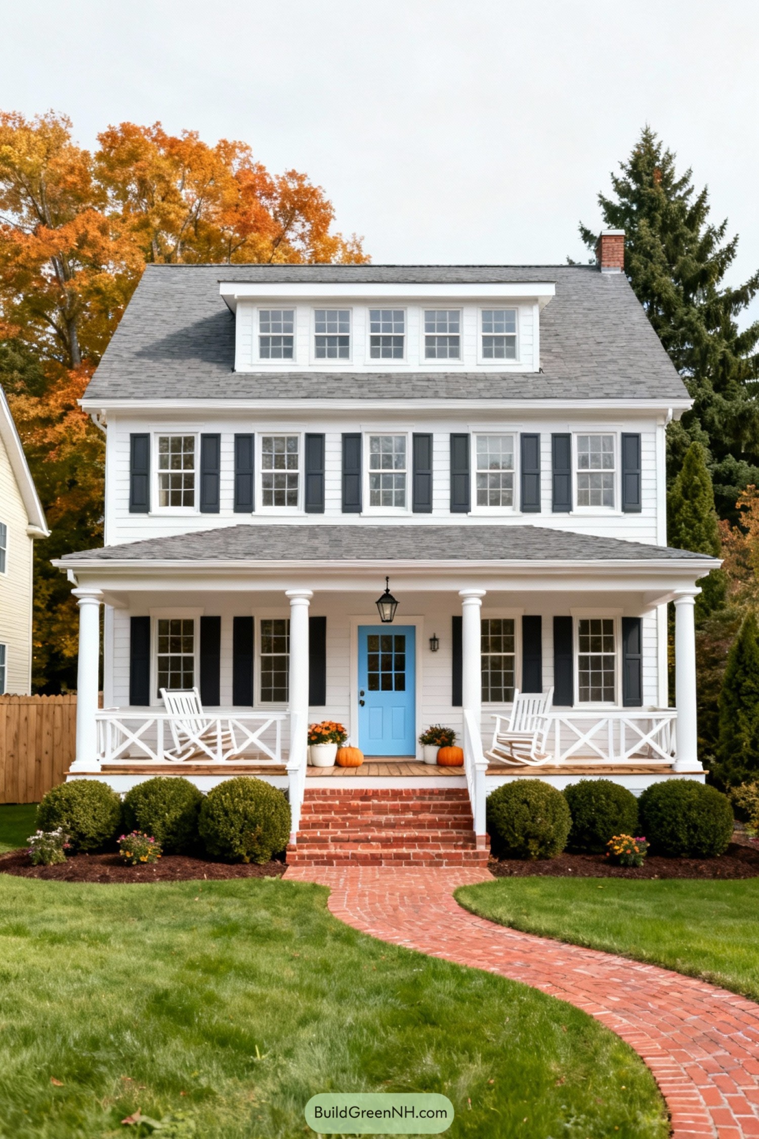 high-res photo of Colonial House With Porch, facade in classic Colonial / Dutch Colonial style with full-width front porch, white horizontal siding, symmetrical front elevation, clean trim lines. Colors: crisp white facade and columns, dark charcoal shutters, medium gray asphalt shingle roof, pale blue front door, warm red brick steps and walkway, green lawn and shrubs, soft autumn foliage tones in background trees. Shape of the structure: two-story rectangular massing with wide front, steep gambrel roof with long shed dormer across upper front, deep roof overhang above porch, balanced window placement across both levels. Materials: painted wood clapboard siding, smooth painted wood trim and porch railings, turned round porch columns, wood porch decking, brick stair risers and walkway, metal porch lantern, asphalt shingles on roof. Roofing: gambrel main roof with front shed dormer, simple white eaves and cornice, minimal ornament, single small chimney barely visible behind ridge. Windows style: evenly spaced double-hung windows with multiple panes, white frames, dark shutters on both levels, three grouped dormer windows centered above, additional singles along dormer, ground floor windows flanking the entry and near porch corners, all set within simple white casings. Door style: single central front door with divided-light upper panel, solid lower panel, painted pale blue, framed by white trim, simple metal hardware, traditional black hanging lantern centered above entry in porch ceiling. Outdoor area: elevated front porch running full width, white balustrades with crisscross X-pattern panels, several white rocking chairs along porch, brick steps centered and flared slightly, decorated with assorted white and orange pumpkins and potted flowering plants on each side of steps. Landscaping: lush, neatly mowed front lawn, curved red brick path leading from foreground to porch steps, dense low hedges and rounded shrubs planted along porch edge, small flowering plants at base of shrubs, mulched planting beds in dark brown soil, mature trees and tall shrubs surrounding property edges. Surrounding background: quiet suburban setting, partial neighboring light-colored house visible at left, wooden privacy fence behind left side yard, tall deciduous trees with early autumn orange and golden leaves mixed with deep green evergreens, soft bright sky with light overcast creating even, gentle daylight. single real-life photo, high-resolution, architectural photography, soft lighting, cinematic composition, strictly no collages