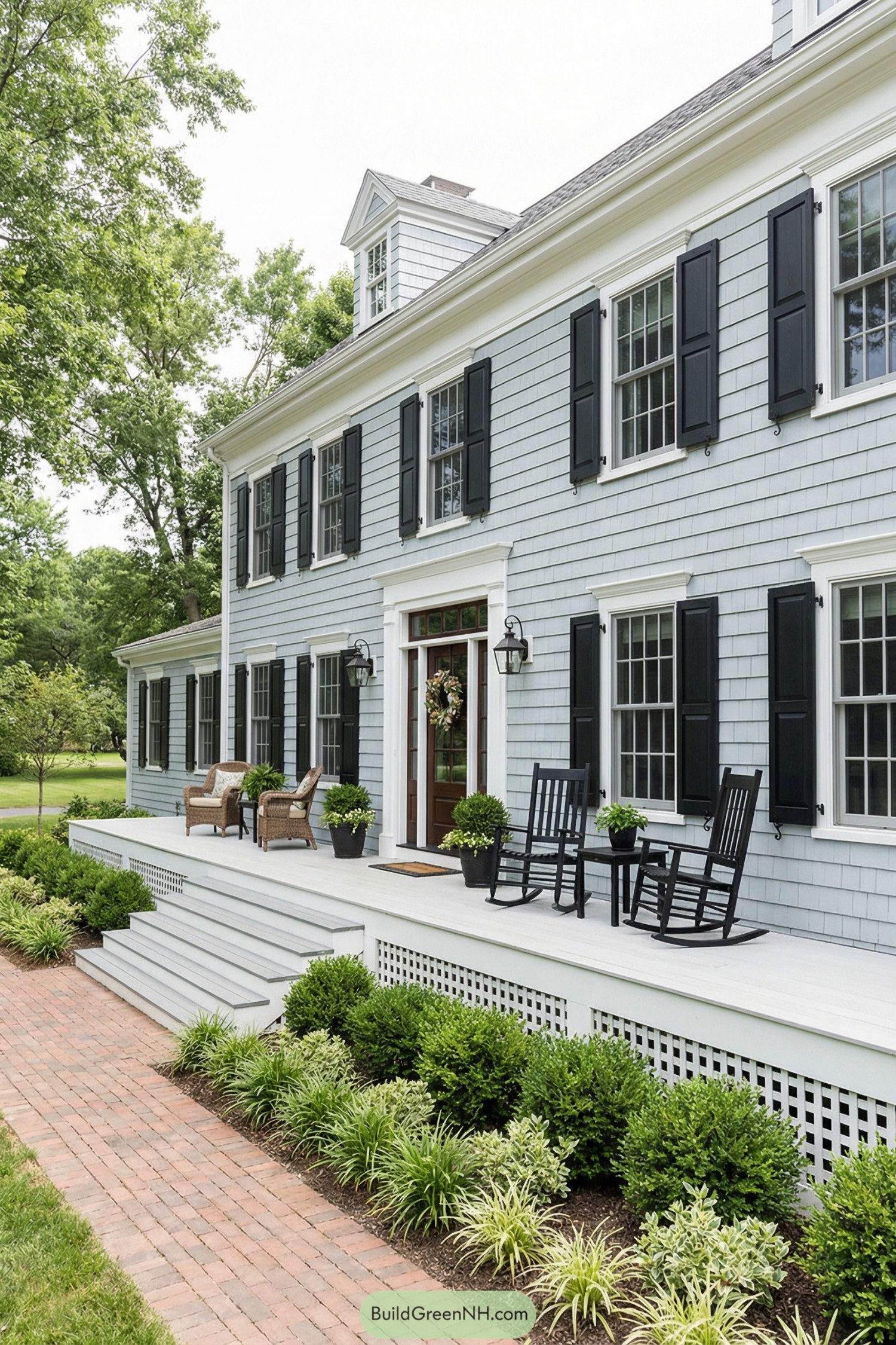 Light blue shingle colonial home with wide white front porch, black shutters, and brick walkway bordered by lush plantings