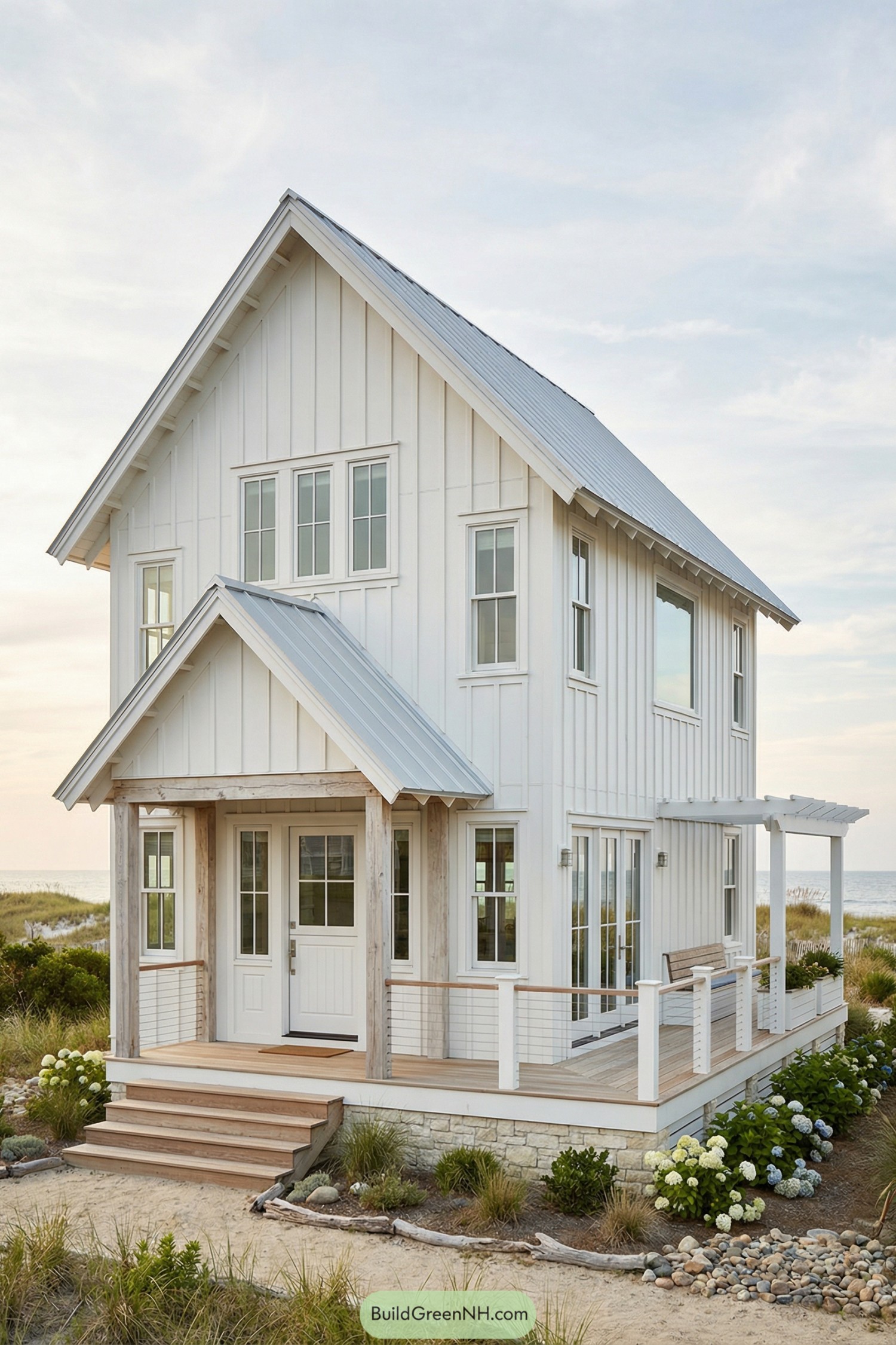Two-story white beach cottage with metal roof and wraparound porch beside sandy dunes and ocean