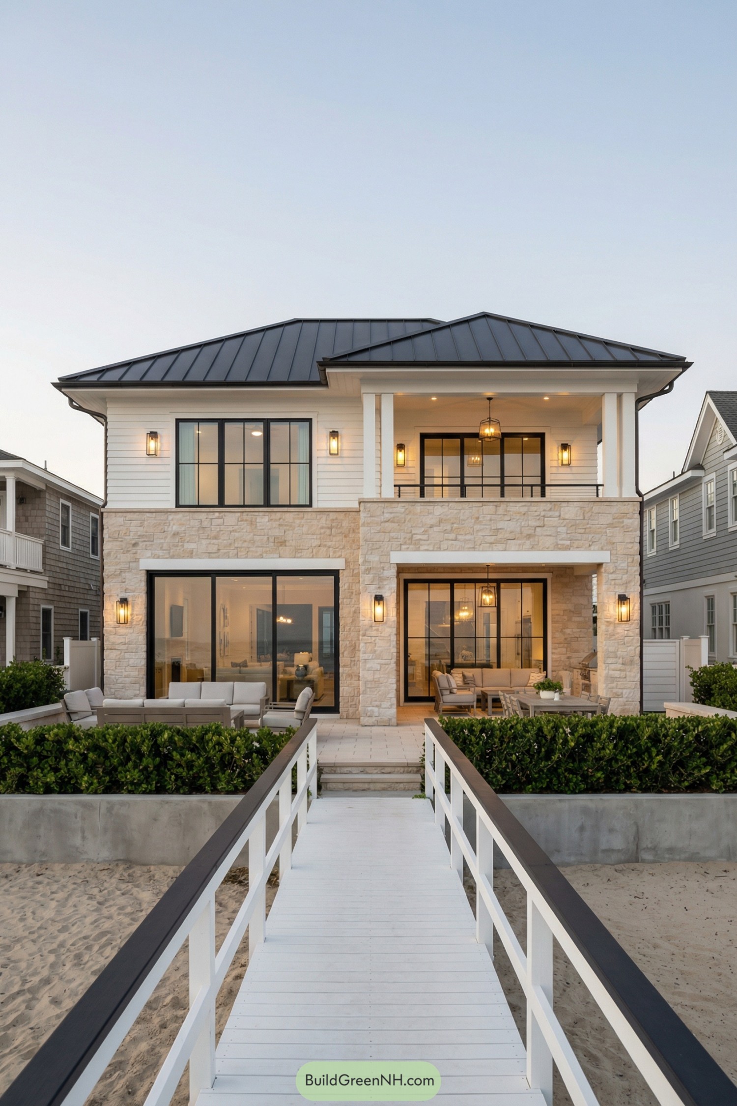 Two-story beach house with stone and white siding, black metal roof, and broad glass doors opening to oceanfront patios and a boardwalk