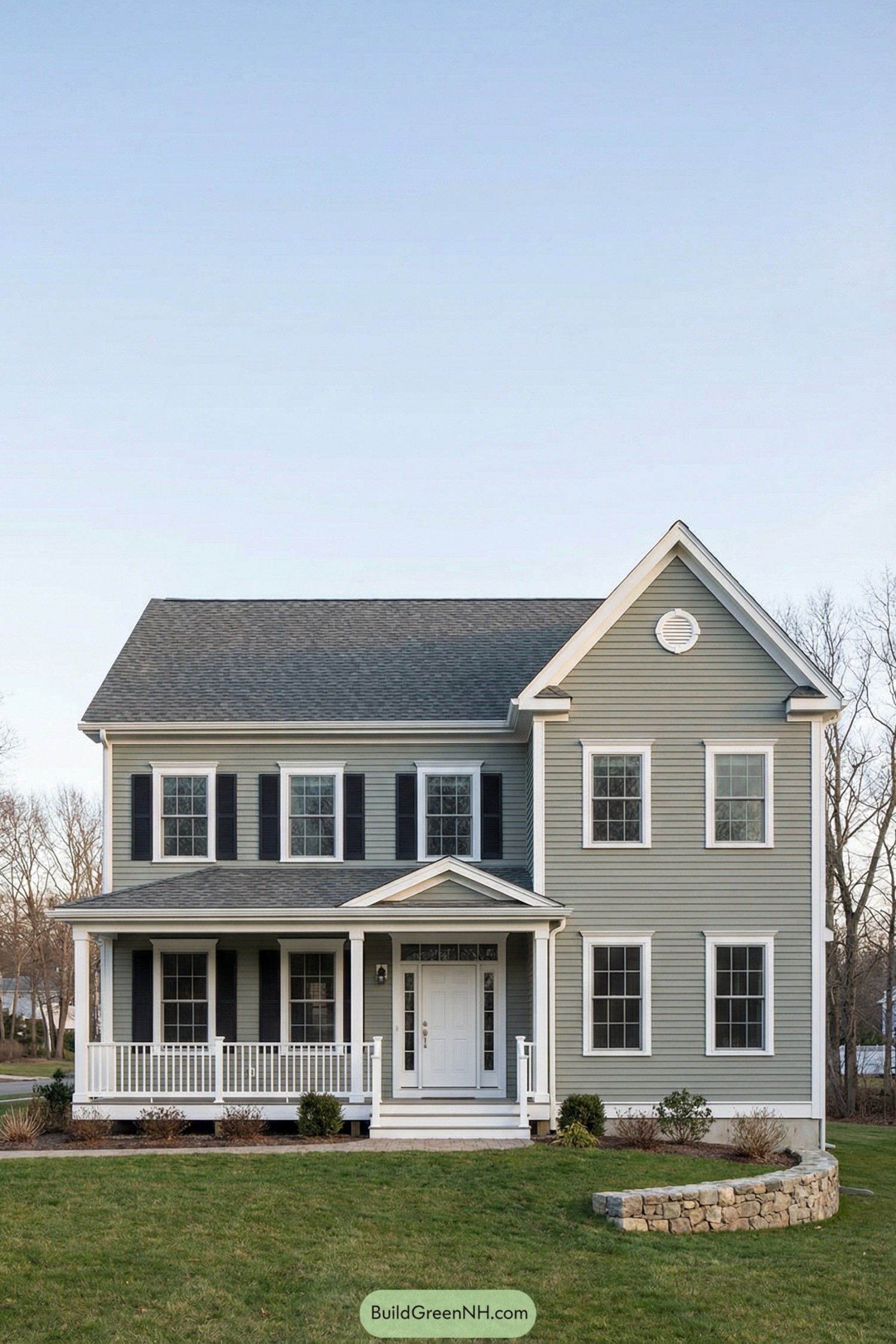 Two story garrison colonial home with sage siding white trim black shutters and a front porch. Stone edging borders a small landscaped garden beside the lawn