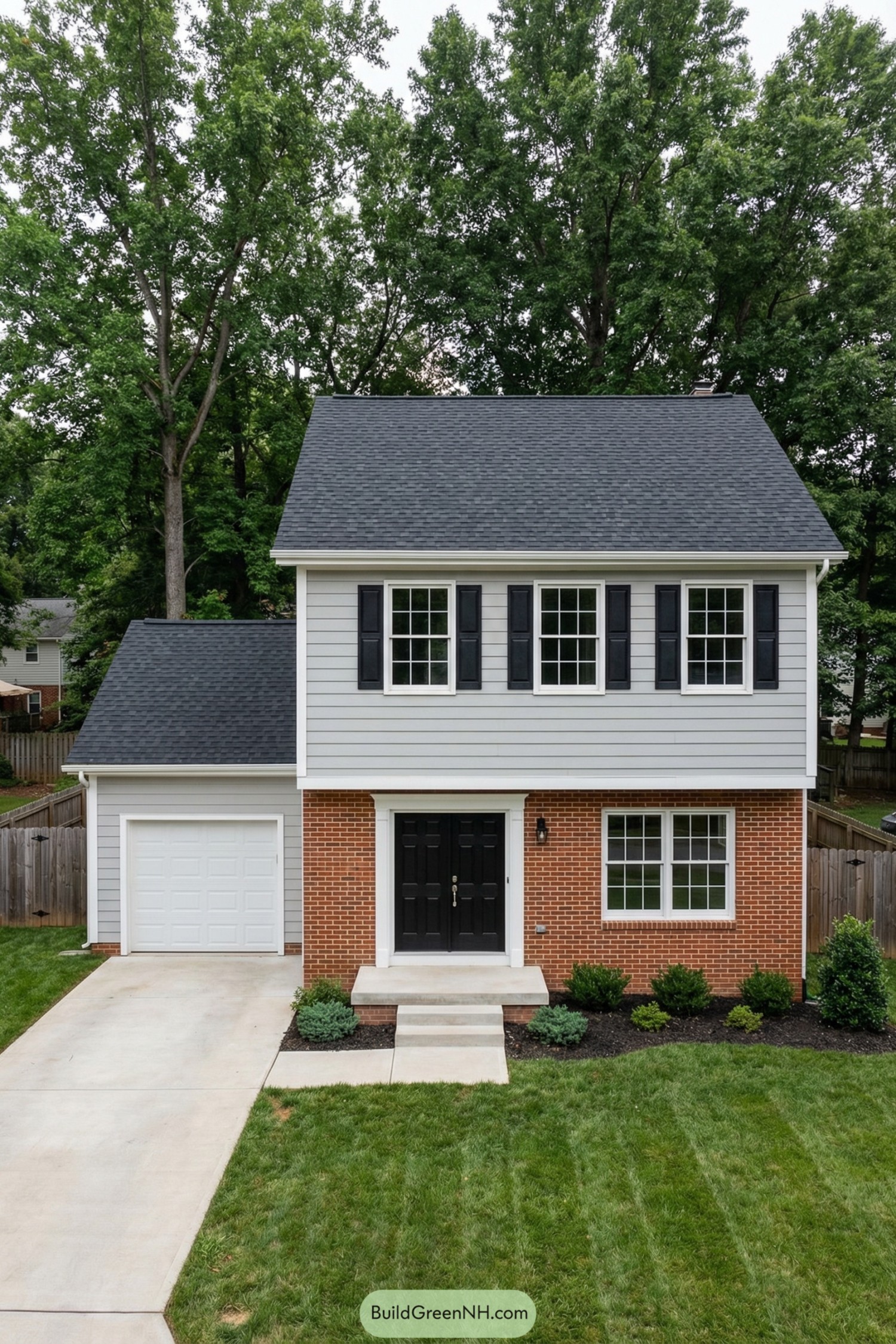 Two story garrison colonial with brick first floor, gray siding above, black shutters, and attached one car garage. Neat front lawn, simple shrubs, and concrete driveway complete the tidy facade