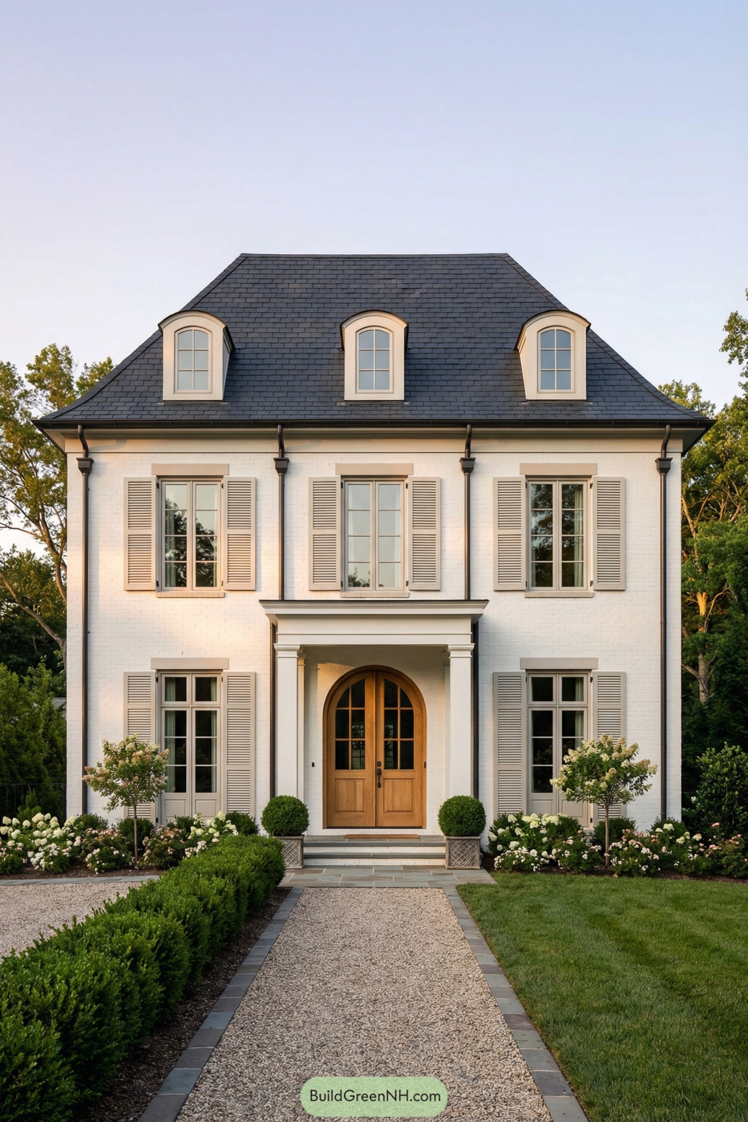 high-res photo of modern french country house exterior, symmetrical facade in refined french country style, painted white brick walls with light warm-gray trim and shutters, tall rectangular massing with two full stories and an attic level under a steep hipped roof, dark charcoal slate-style roof with three evenly spaced arched dormer windows, slender black metal gutters and downspouts, large vertically proportioned french casement windows with divided lights and soft-gray shutters, central arched double wooden entry door with multiple glass panes and warm natural wood tone, shallow front portico with two square white columns and simple entablature, small stone stoop with a few steps framed by white box planters holding clipped round topiary shrubs, straight central gravel walkway edged with dark stone pavers leading from foreground to entrance, low manicured boxwood hedges bordering the path and flower beds, neatly edged deep-green lawn in the foreground, beds with white and blush flowering shrubs and small ornamental trees on both sides of the facade, dense mature greenery and trees framing the property in the background, clear sky with soft gradient and gentle warm evening light enhancing shadows and depth, single real-life photo, high-resolution, architectural photography, soft lighting, cinematic composition, strictly no collages.