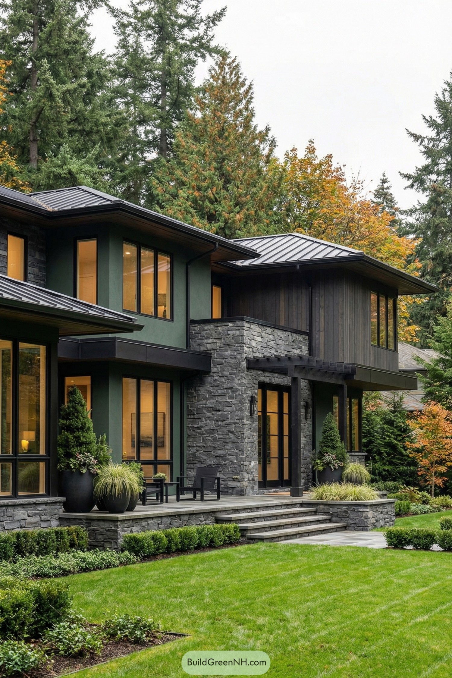 Contemporary two story house with dark green walls stone accents and large windows opening to a manicured lawn and tall forest backdrop